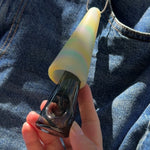 Hand holding a glass pipe with a pastel rainbow silicone cover, clipped to denim fabric in sunlight.