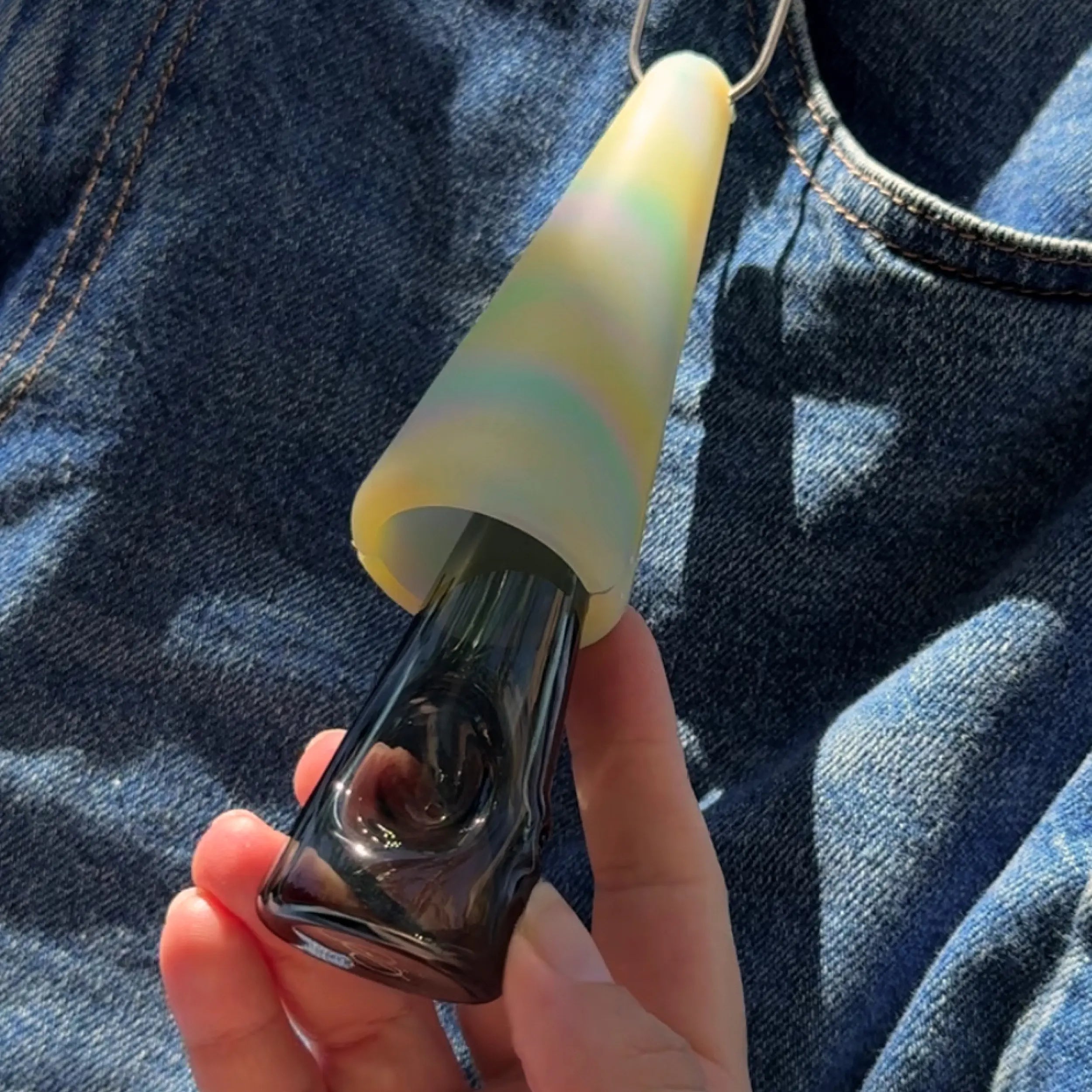 Hand holding a glass pipe with a pastel rainbow silicone cover, clipped to denim fabric in sunlight.
