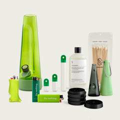 Session Goods Designer Series Ultimate Bundle in green tones, featuring a translucent green bong, matching silicone accessories, two ‘Do nothing.’ branded lighters, a clear water bottle labeled ‘Dry Mouth Erase.’, a sleek one-hitter pipe with cone sleeve, a black four-piece grinder, and two stash pods with green caps—all arranged on a neutral background.