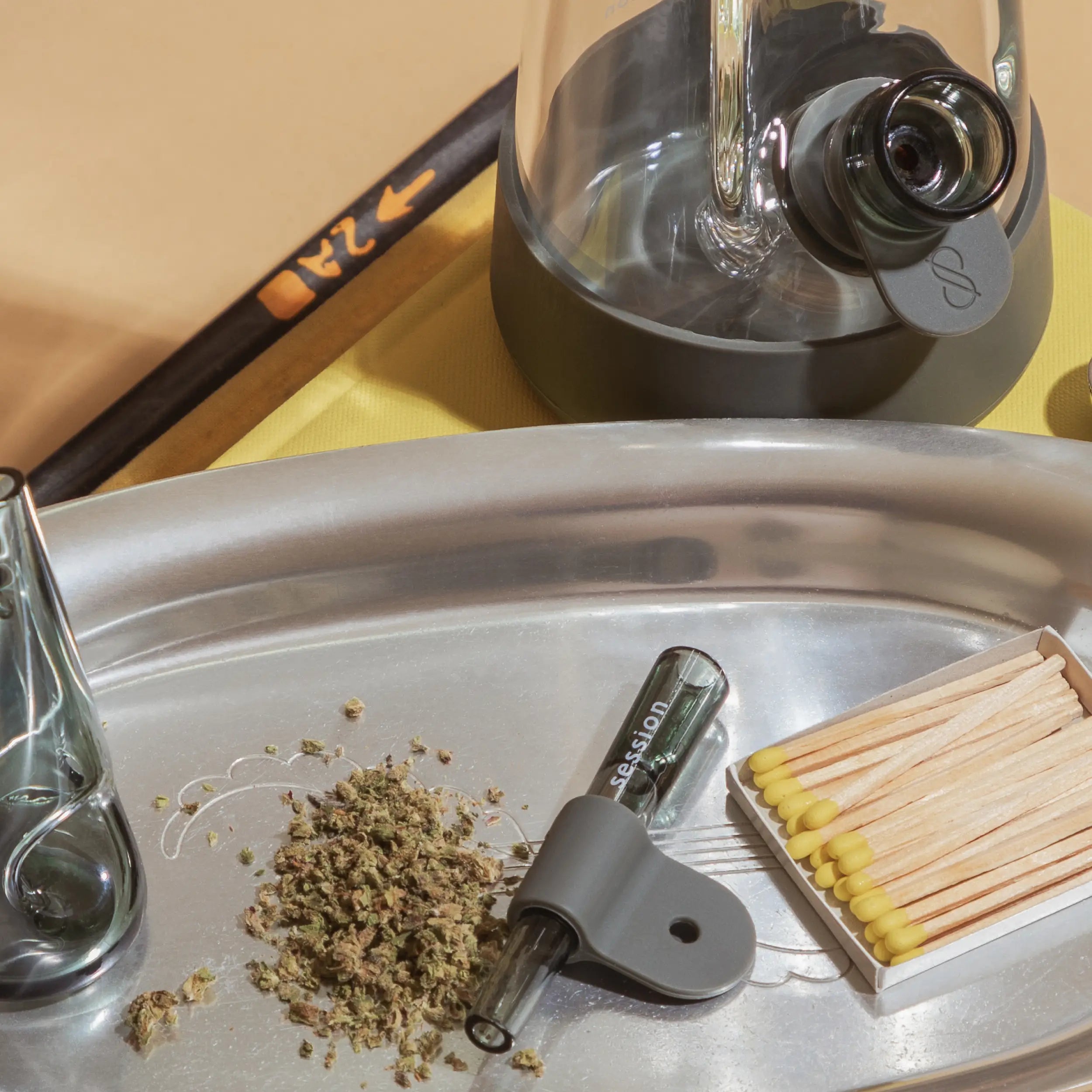 Charcoal one hitter on tray next to grounded herb, with a session goods portable pipe and bong all in charcoal color