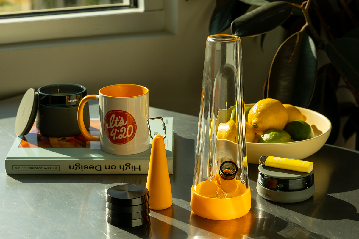 Glass bong with yellow base styled on a table with citrus, a coffee mug, and design books, bright fall lifestyle reset scene.