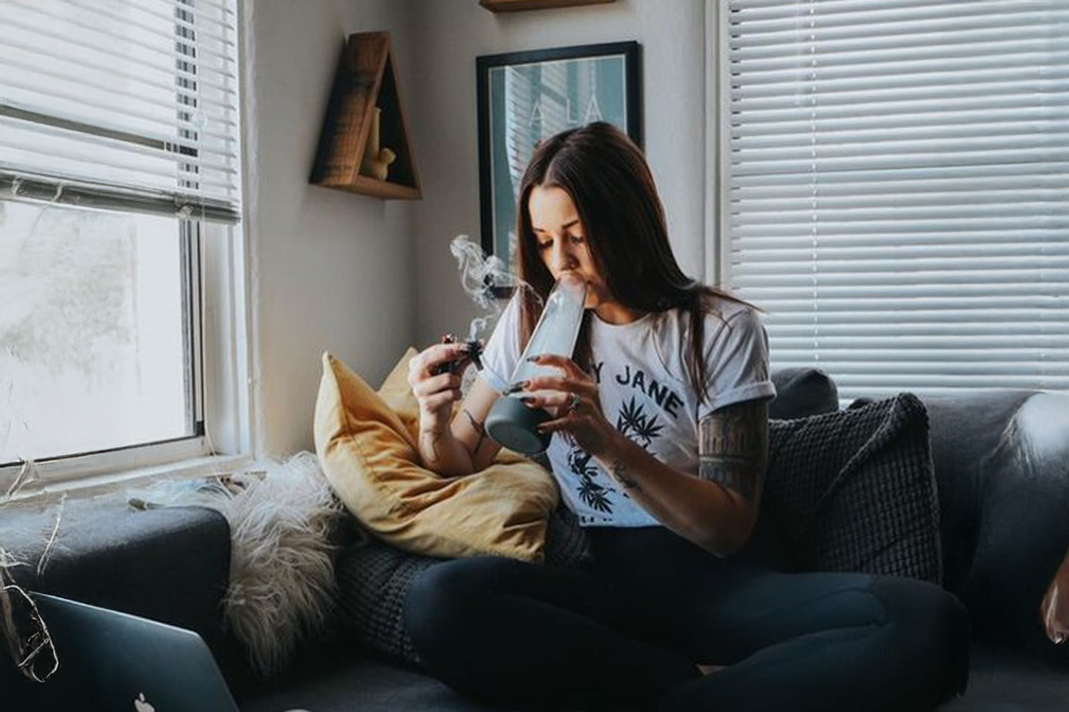 A woman mid-inhale with her bong on a couch in comfy clothes