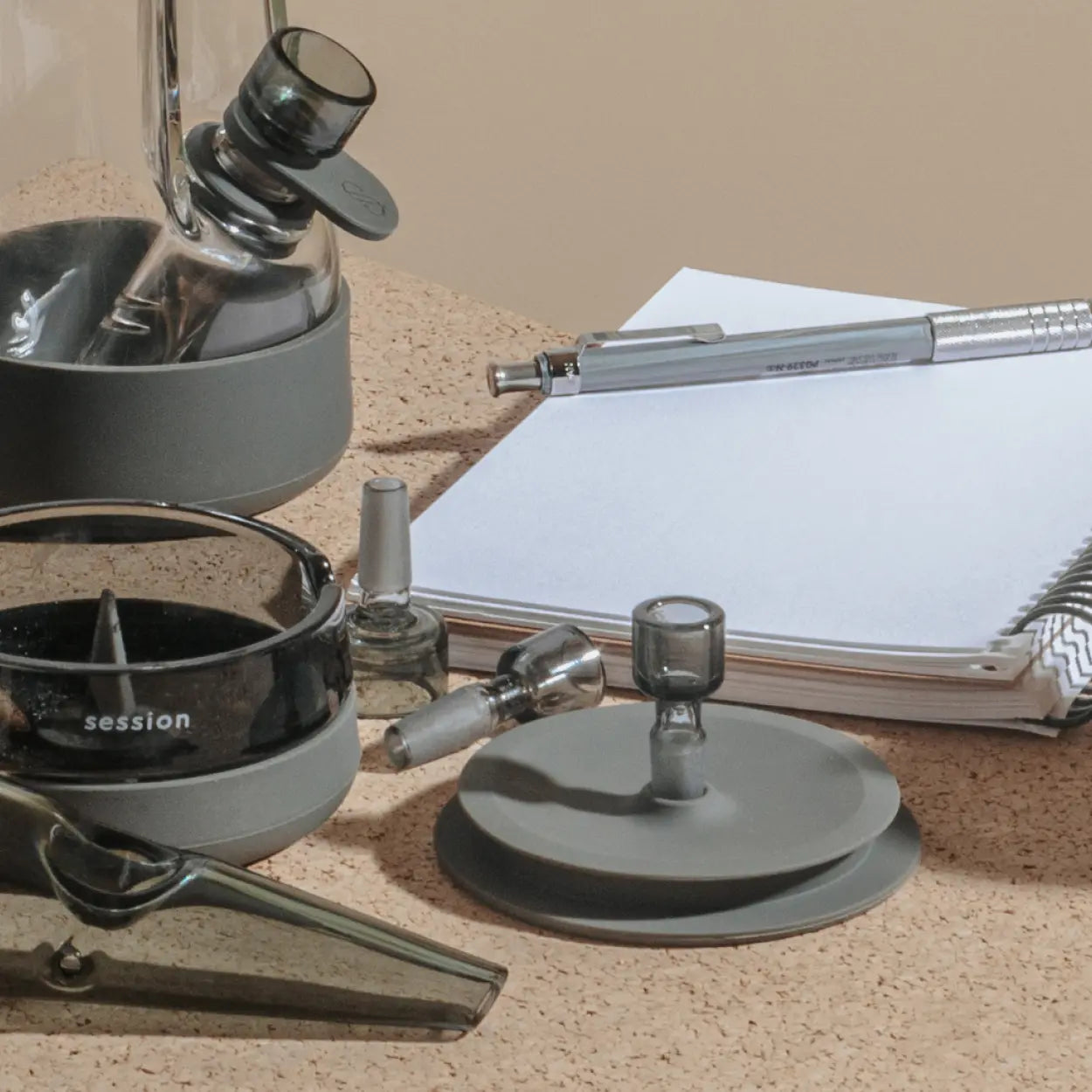 Collection of Session Goods smoking accessories in grey, including a bong base, ashtray, bowl pieces, downstems, and a pipe, arranged on a cork surface beside a silver pen and a spiral notebook