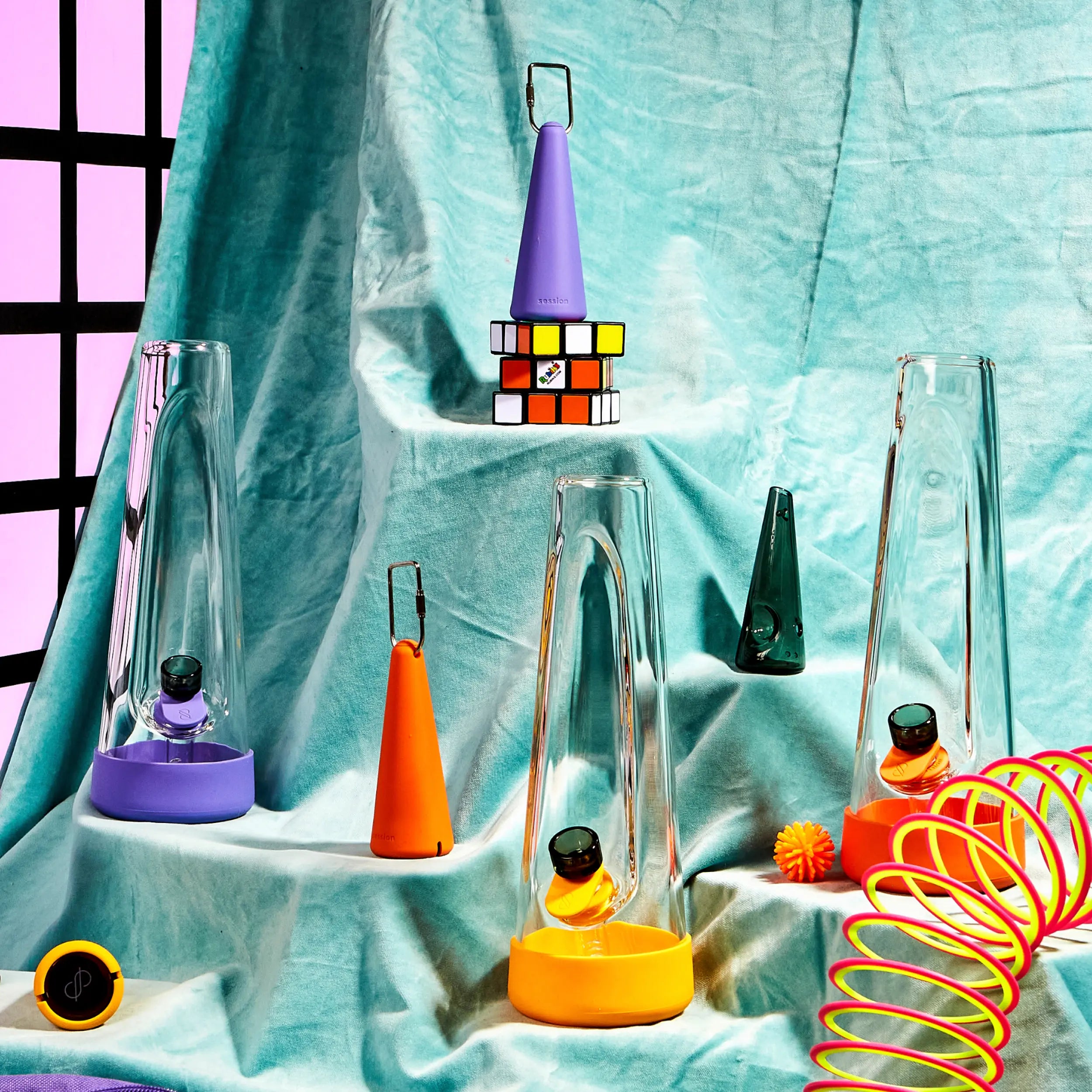 A close-up of Session Goods bongs in vibrant silicone bases—purple and orange—arranged with colorful props like a Rubik’s Cube, slinky, and retro toys, all set on soft teal velvet with a bold grid backdrop.