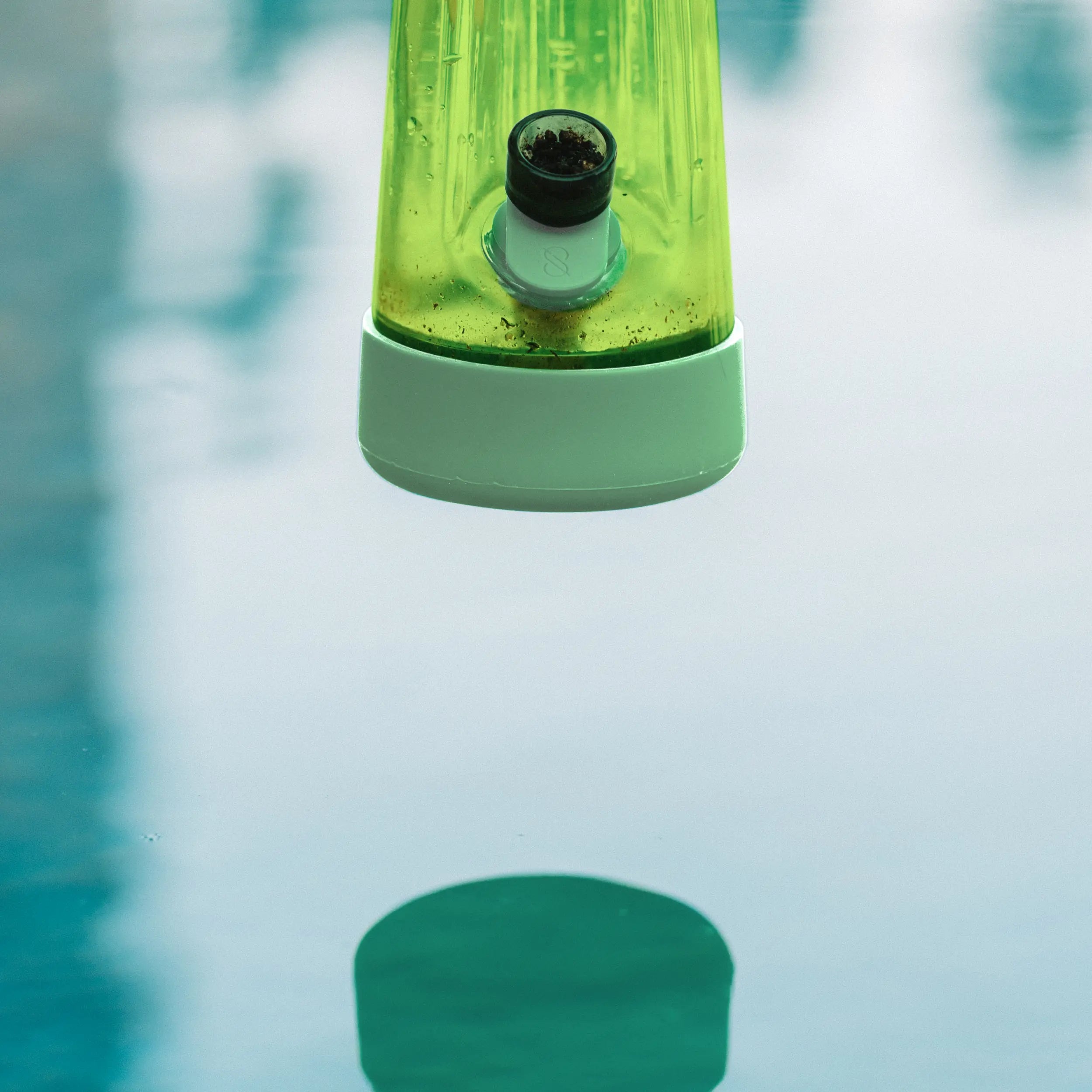 Close-up of a green Session Goods Designer Series bong Grass hovering over a pool, with its reflection visible in the water below