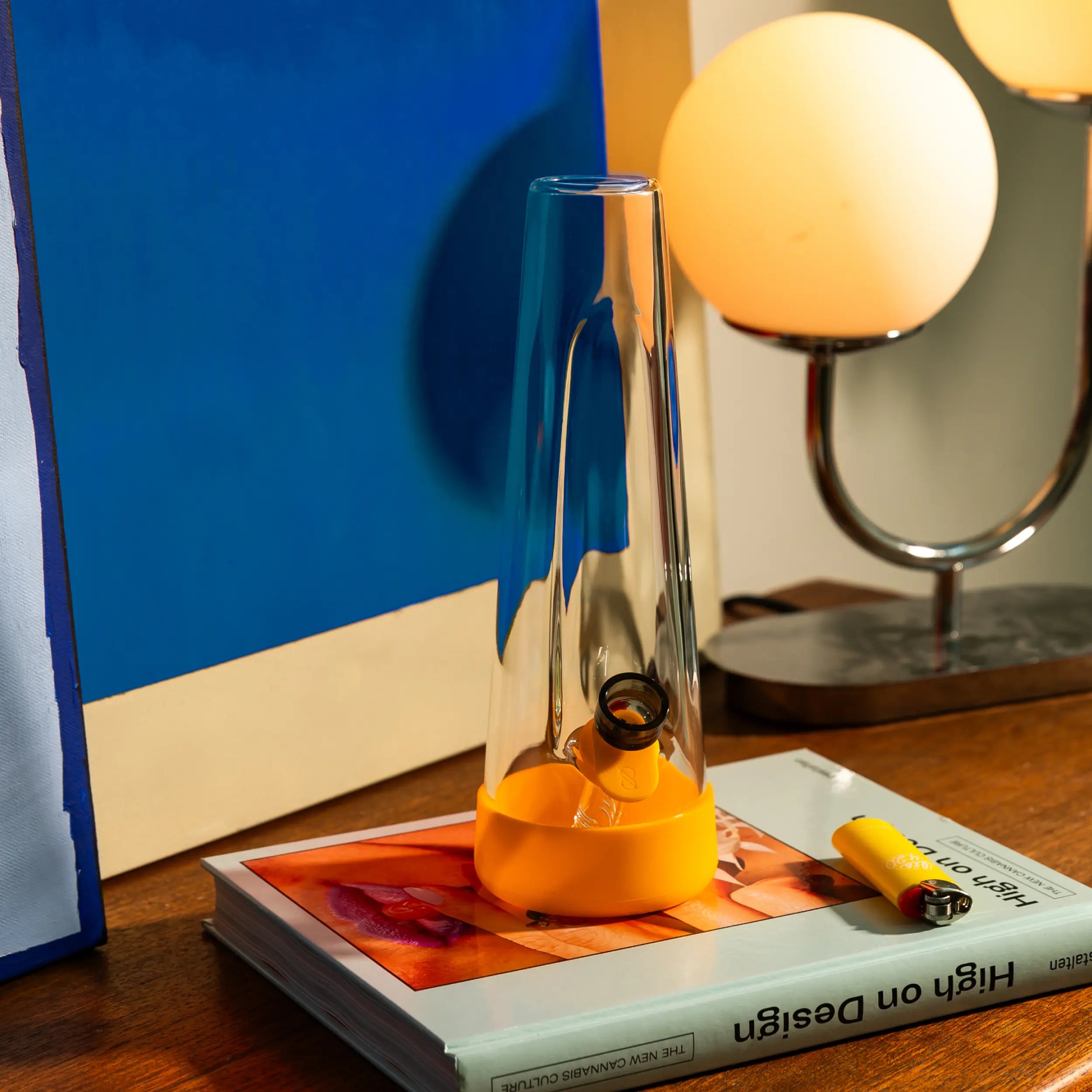 Close-up of a Session Goods tapered glass bong with a yellow silicone base, sitting on a "High on Design" book next to a matching yellow lighter. Vertical mobile homepage banner.
