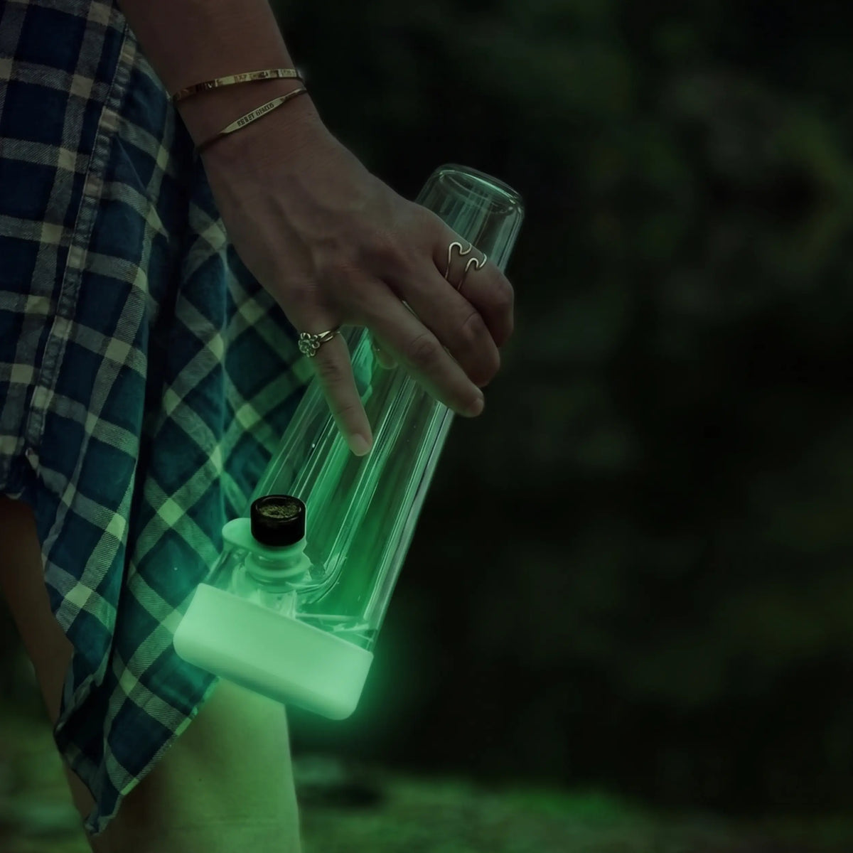 Person holding a Session Goods clear glass bong with a green glow-in-the-dark silicone base while wearing a blue flannel shirt.