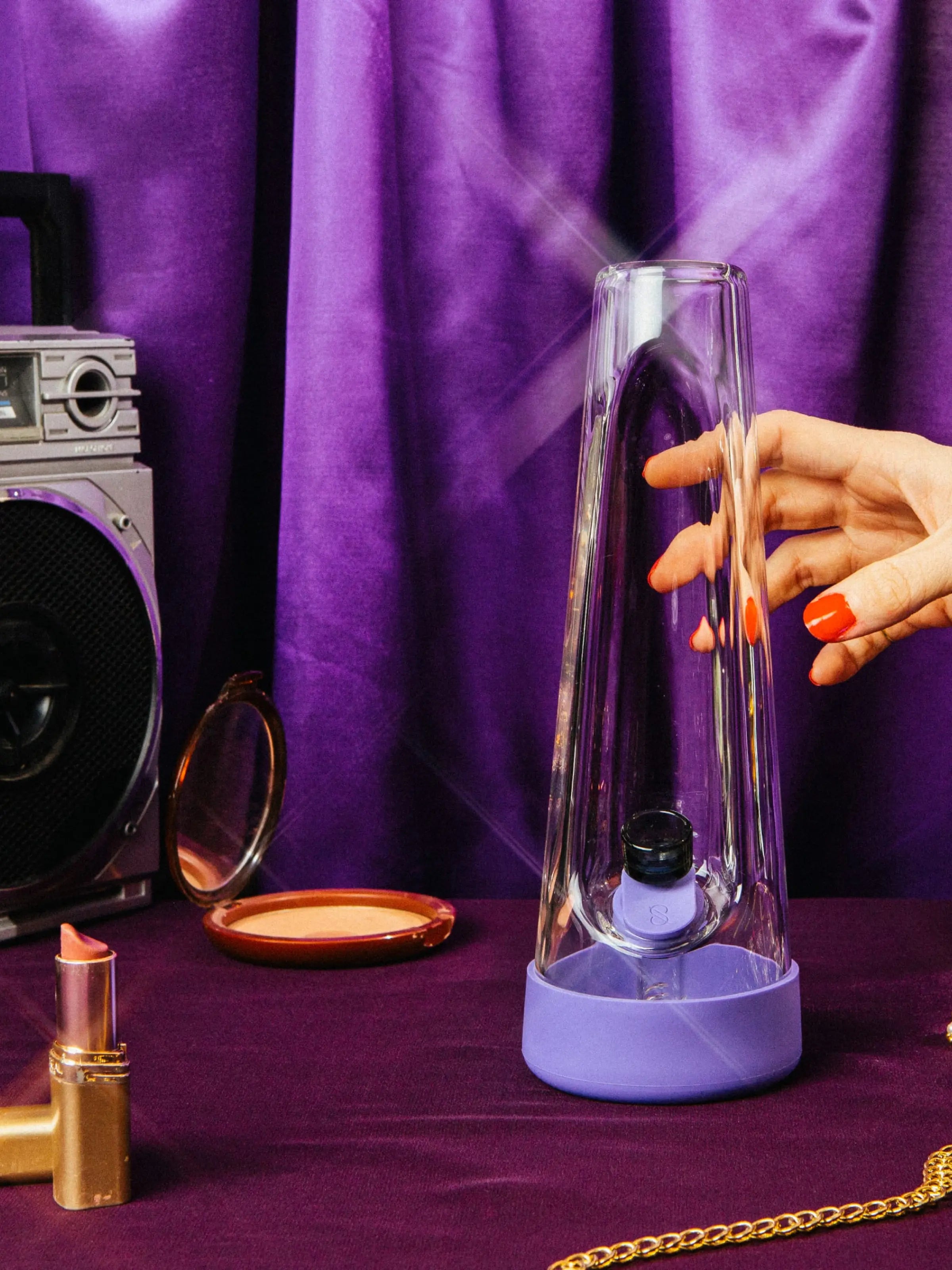 An aesthetic, 70s-inspired setup featuring the moonlight purple Session Goods water pipe. The sleek glass piece is surrounded by vintage-style accessories, including a classic radio and gold chains, all set against deep purple velvet. The lighting is soft and nostalgic with subtle lens flares, highlighting the modern design of the piece.