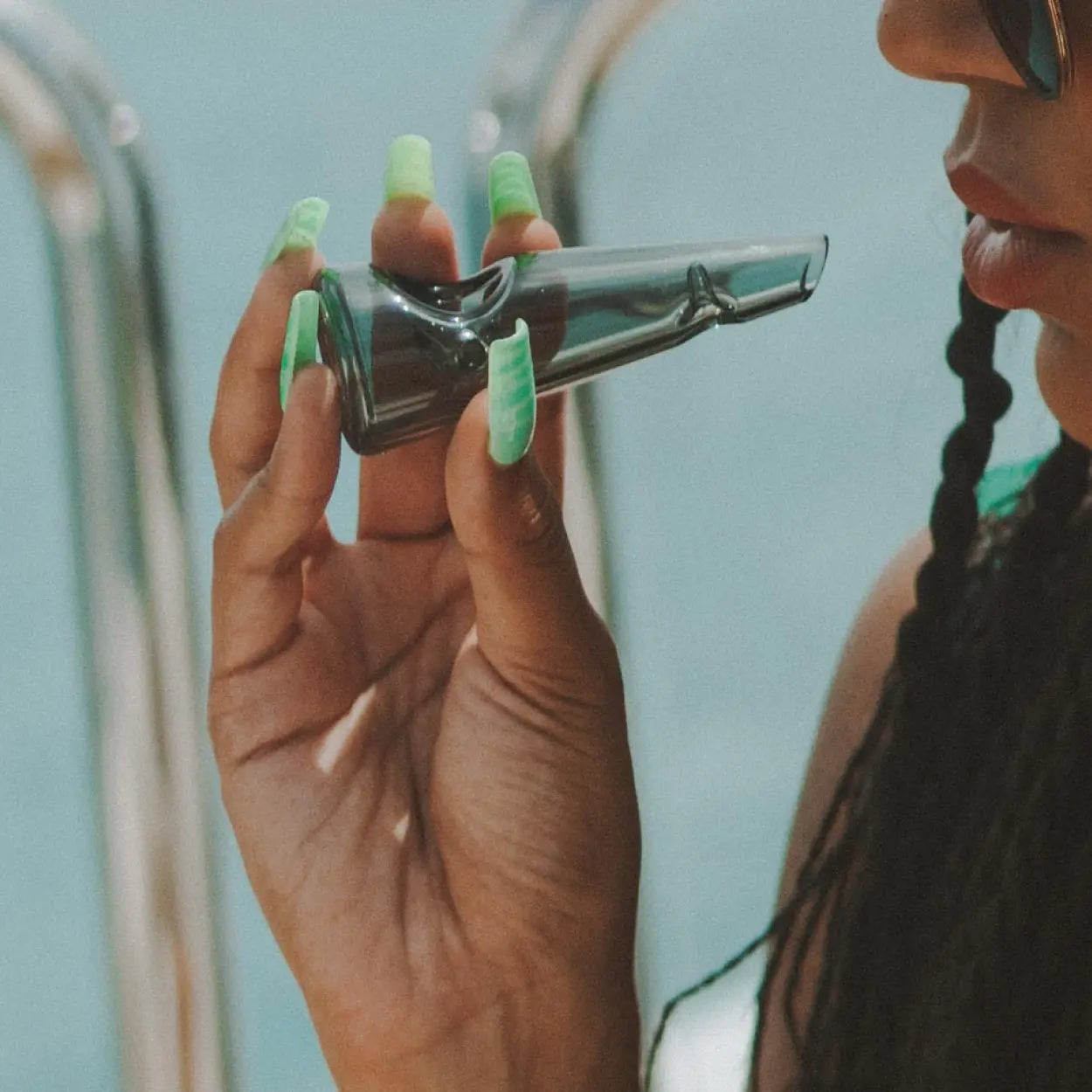 A person with braided hair and lime green nails holds a translucent grey glass hand pipe. The background shows a soft-focus pool ladder and blue water, suggesting a relaxed outdoor setting.
