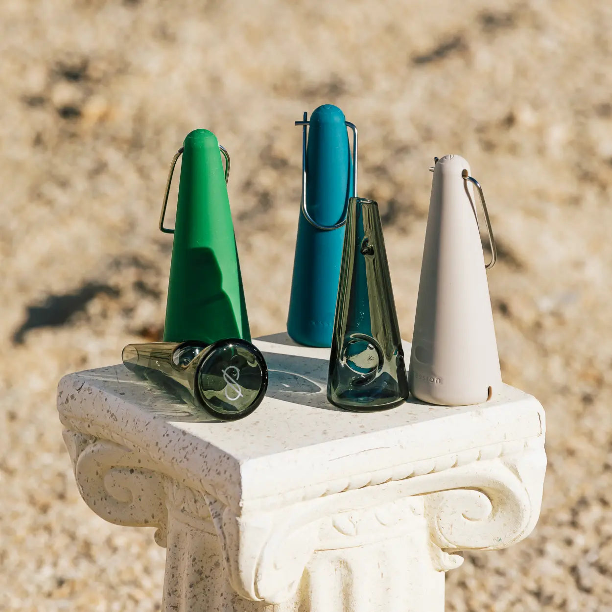 A group of Session Goods pipes and their protective silicone carry cases arranged on a white, ornate stone pedestal. The collection includes a transparent dark glass pipe alongside cases in vibrant green, deep teal, and soft cream. The scene is set outdoors in bright sunlight against a soft-focus, sandy background.