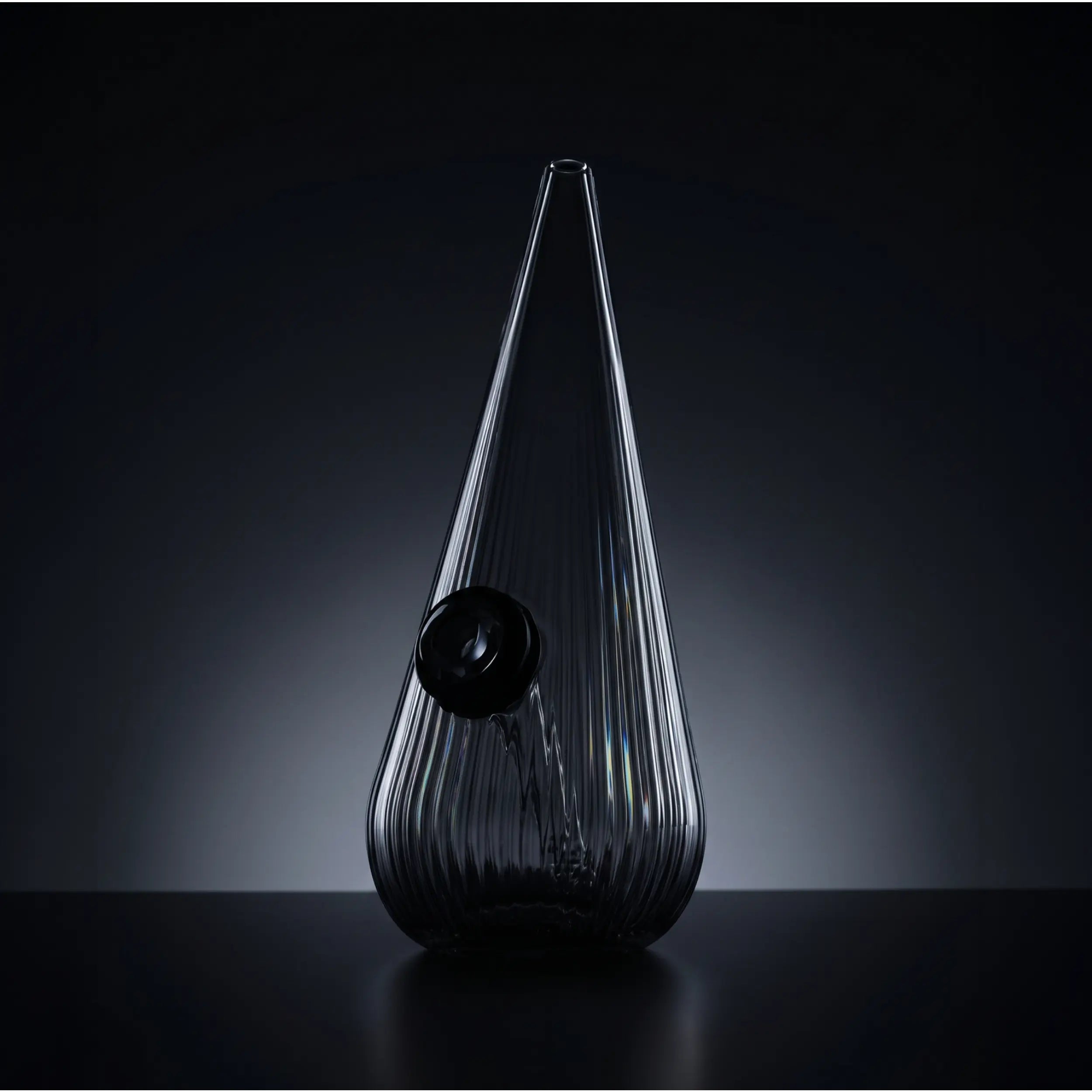 A close-up, vertical shot of a teardrop-shaped ribbed glass bong standing on a reflective black table. Illuminated by moody studio backlight against a dark gradient backdrop, accentuating the vertical ridges and glass contours in a minimalist, high-end product style.