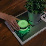 A lifestyle shot of the Session Goods glow-in-the-dark green silicone ashtray on a wooden coffee table. A hand with a ring gently rests near the compact, closed ashtray, which is emitting a strong neon green glow. The ashtray sits on top of an open magazine next to a small green plant in a gray pot.