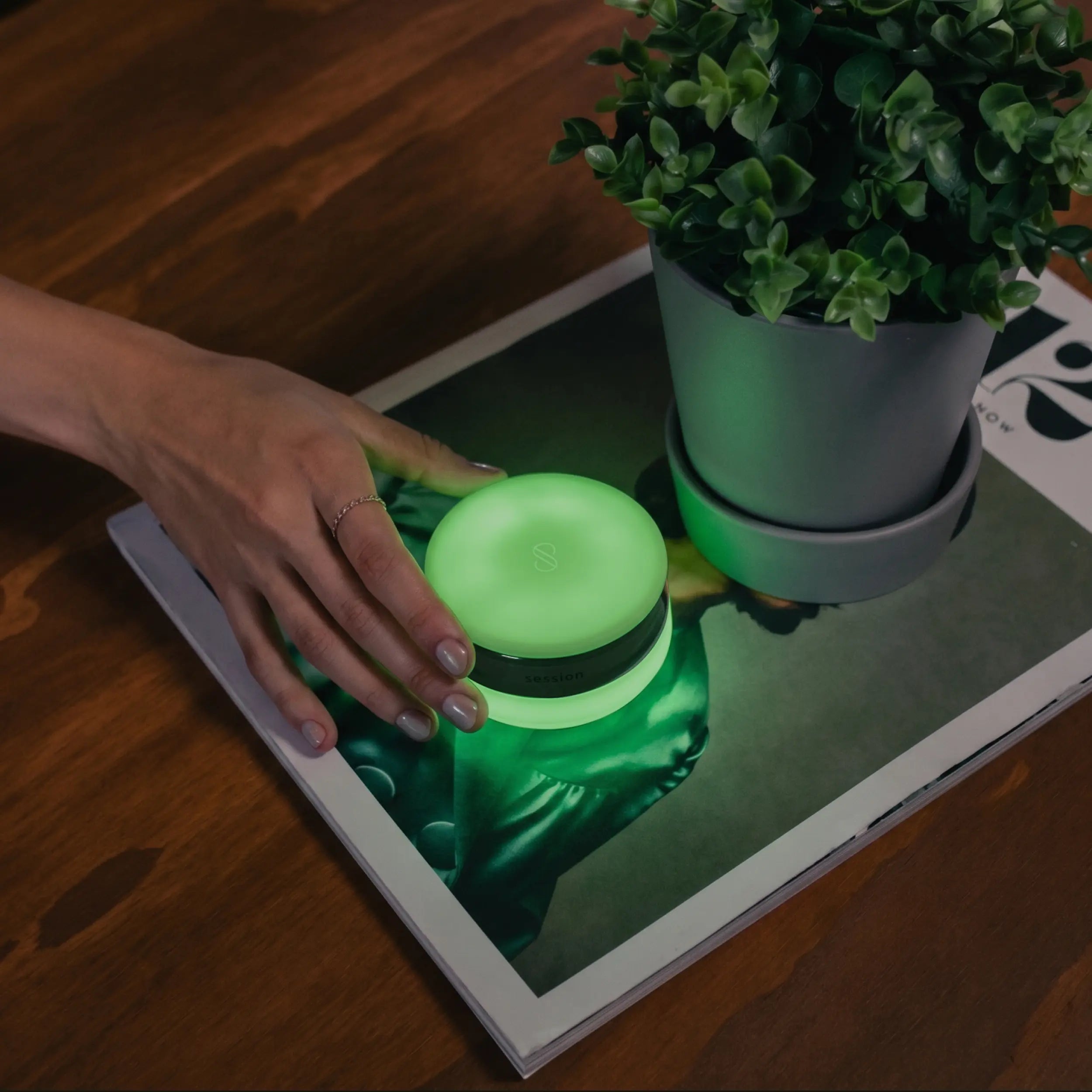 A lifestyle shot of the Session Goods glow-in-the-dark green silicone ashtray on a wooden coffee table. A hand with a ring gently rests near the compact, closed ashtray, which is emitting a strong neon green glow. The ashtray sits on top of an open magazine next to a small green plant in a gray pot.