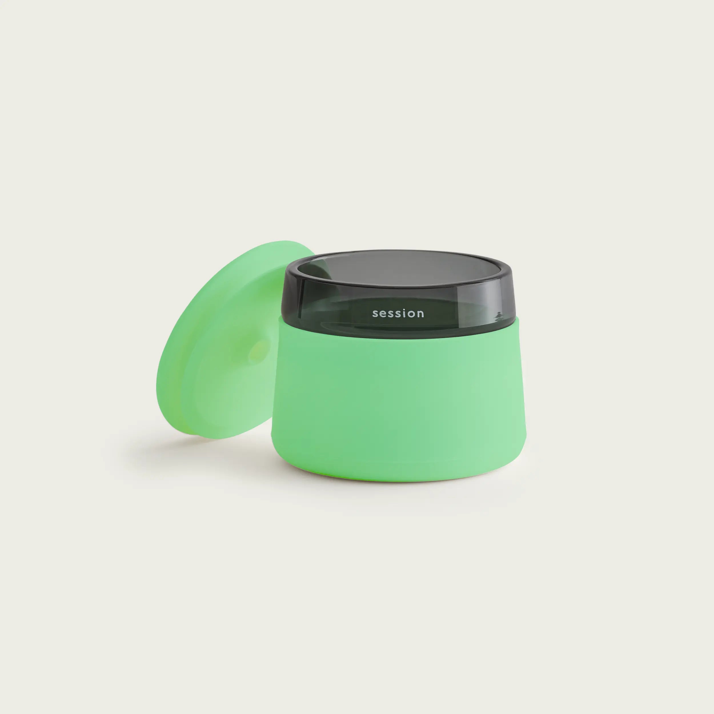 Studio shot of the Session Goods glow-in-the-dark lime green stash jar and ashtray on a white background, with its silicone lid resting against the black glass and green silicone body.