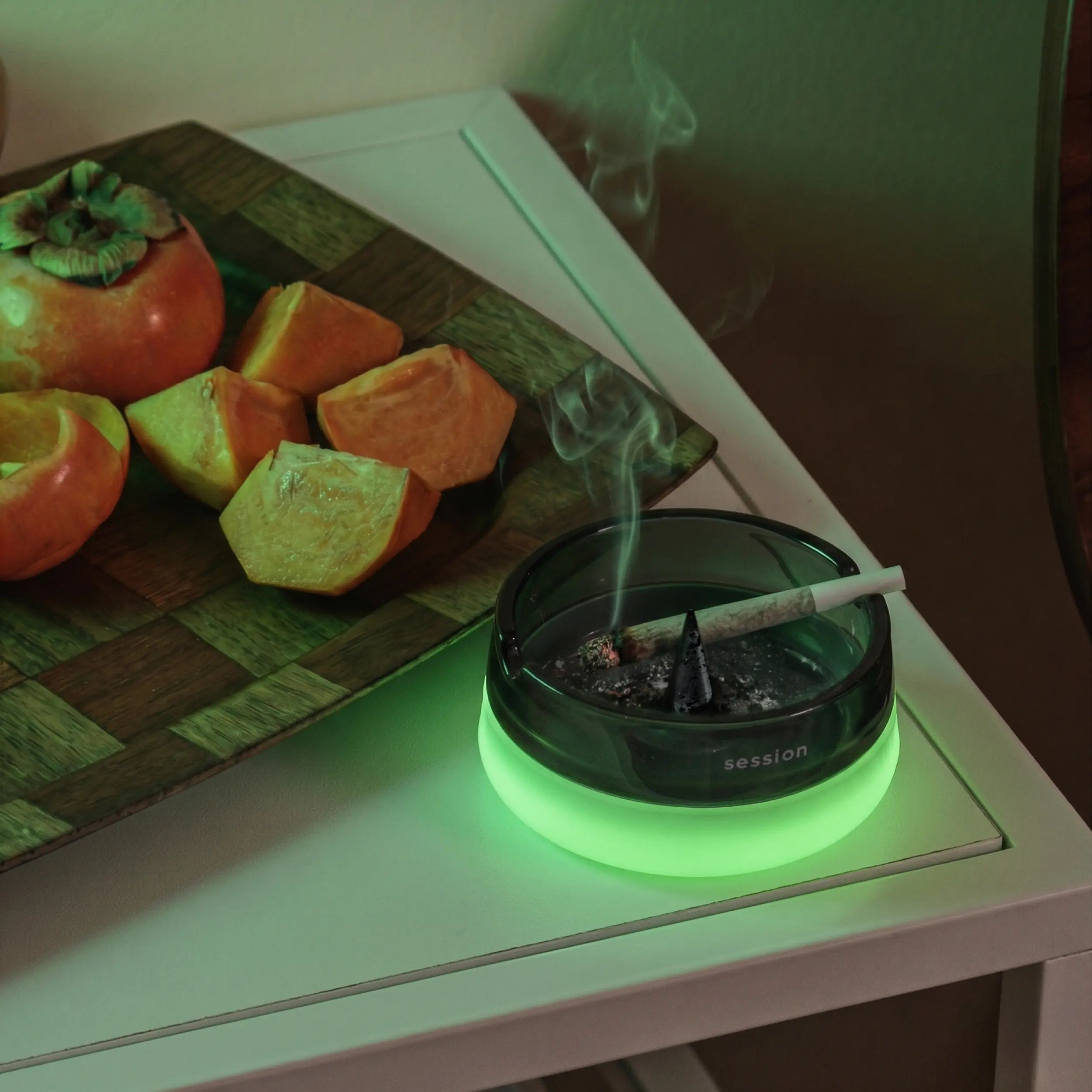 A close-up lifestyle image of an open Session Goods ashtray sitting on a white side table. The dark, smokey glass dish has a glowing green silicone base and a center de-bowler spike. A lit pre-roll rests on the edge with a trail of smoke rising. In the background, a wooden checkered platter holds sliced orange persimmons.