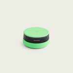 A clean studio product shot against an off-white background showing the Session Goods ashtray in its closed state during daylight. The design features a vibrant, light-green silicone lid and base cradling a translucent, dark-gray tinted glass body with the word "session" printed on the side.