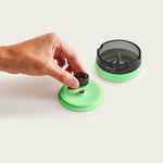 A studio shot on a plain background showing the modular components of the Session Goods green ashtray. A hand places a dark glass bowl piece onto the inverted light-green silicone lid, demonstrating its dual use as a bowl stand. Next to it sits the main dark tinted glass ashtray with its built-in center cleaning spike and green silicone base.