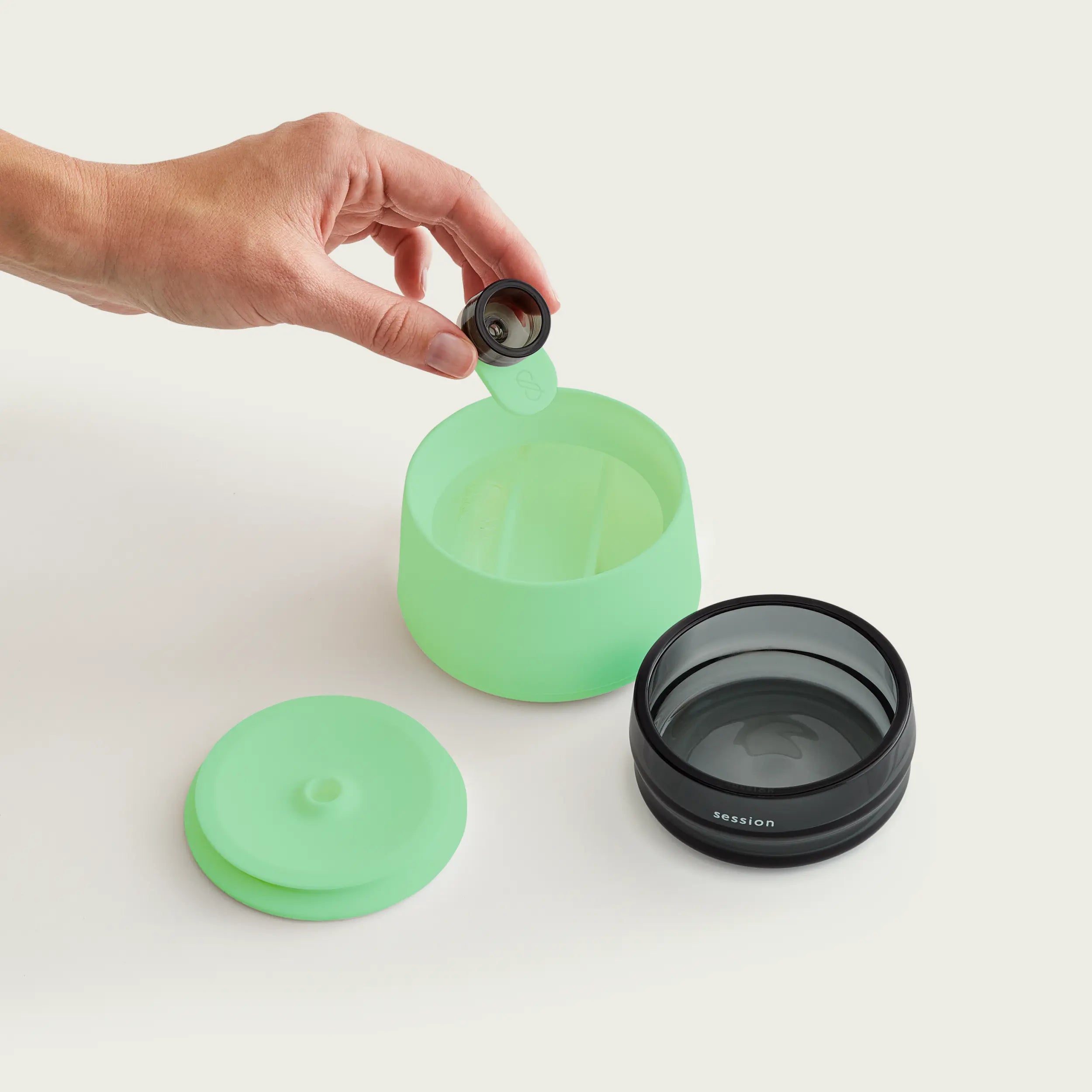 Deconstructed view of the Session Goods glow-in-the-dark stash jar, displaying the lime green silicone base, black glass ashtray insert, silicone lid, and a hand holding a glass bowl piece with a green silicone debowler tool.