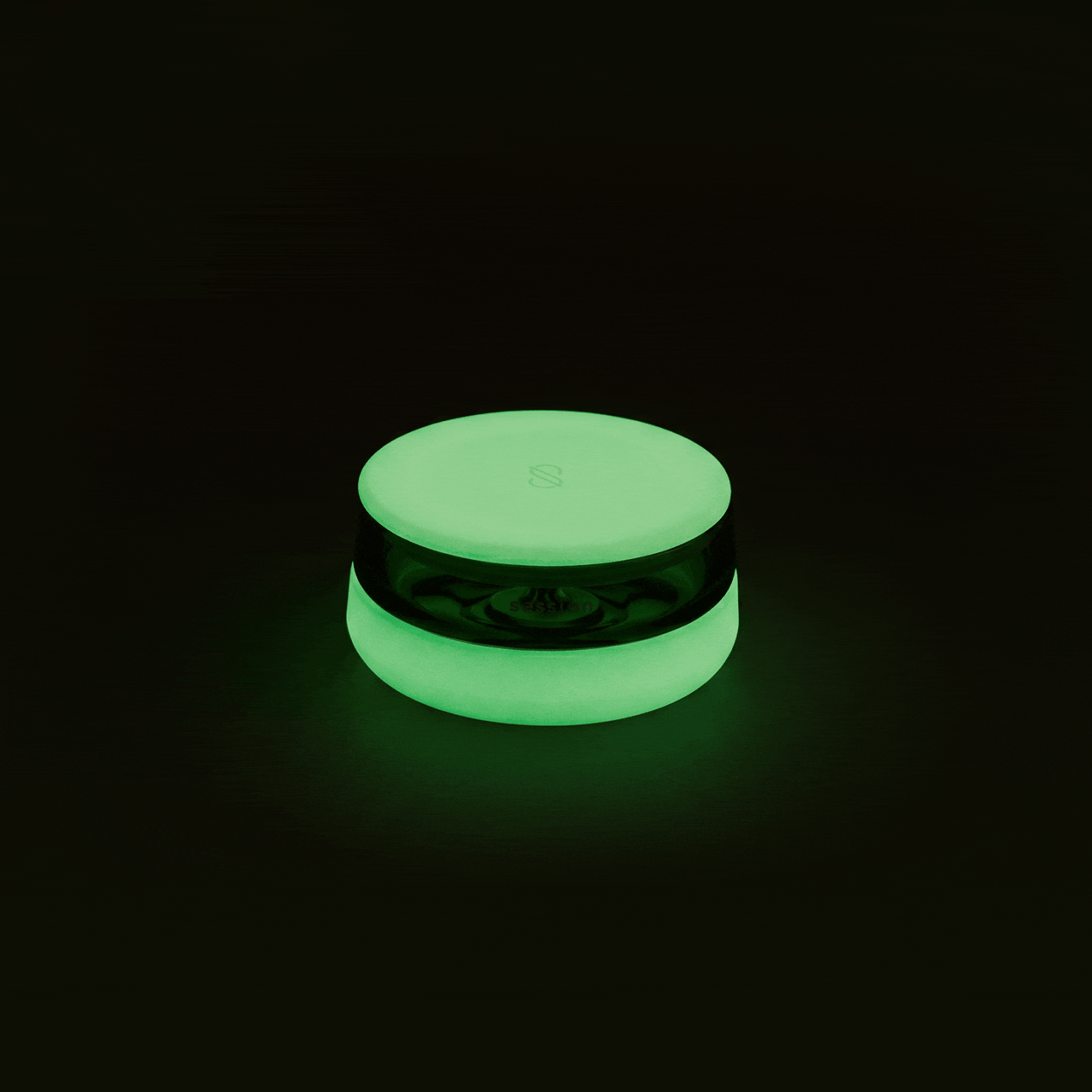 A studio product shot showing the Session Goods circular silicone and glass ashtray completely illuminated with a vibrant, phosphorescent neon green glow centered against a stark black background.