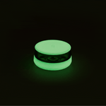 A studio product shot showing the Session Goods circular silicone and glass ashtray completely illuminated with a vibrant, phosphorescent neon green glow centered against a stark black background.