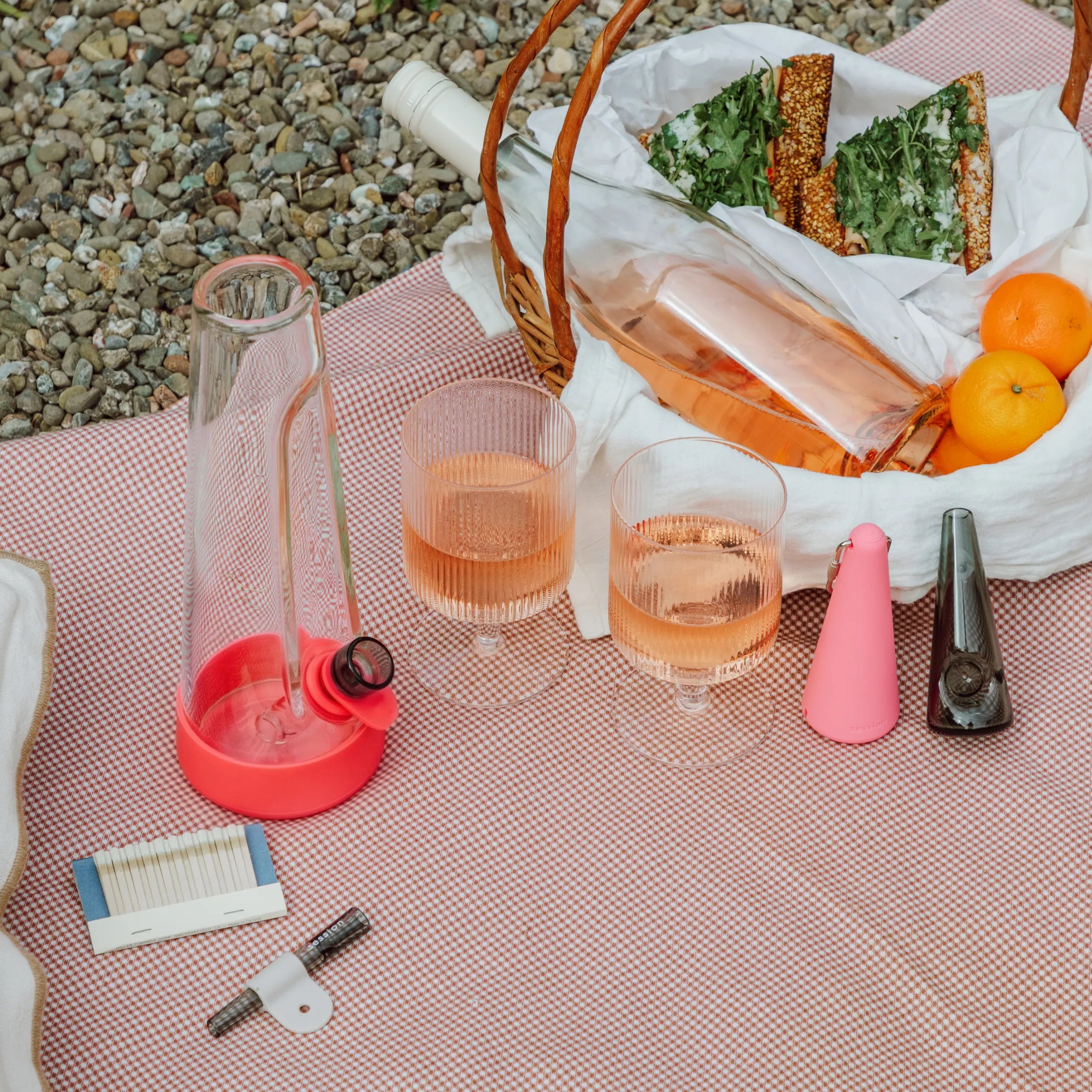 A stylish outdoor picnic setup on a red and white checkered blanket featuring a Session Goods glass bong with cherry red silicone, glasses of rosé wine, sandwiches, fruit, and modern smoking accessories.