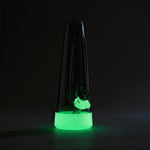 A conical dark glass water pipe with a mint green silicone base and bowl ring glowing brightly in a dark room.