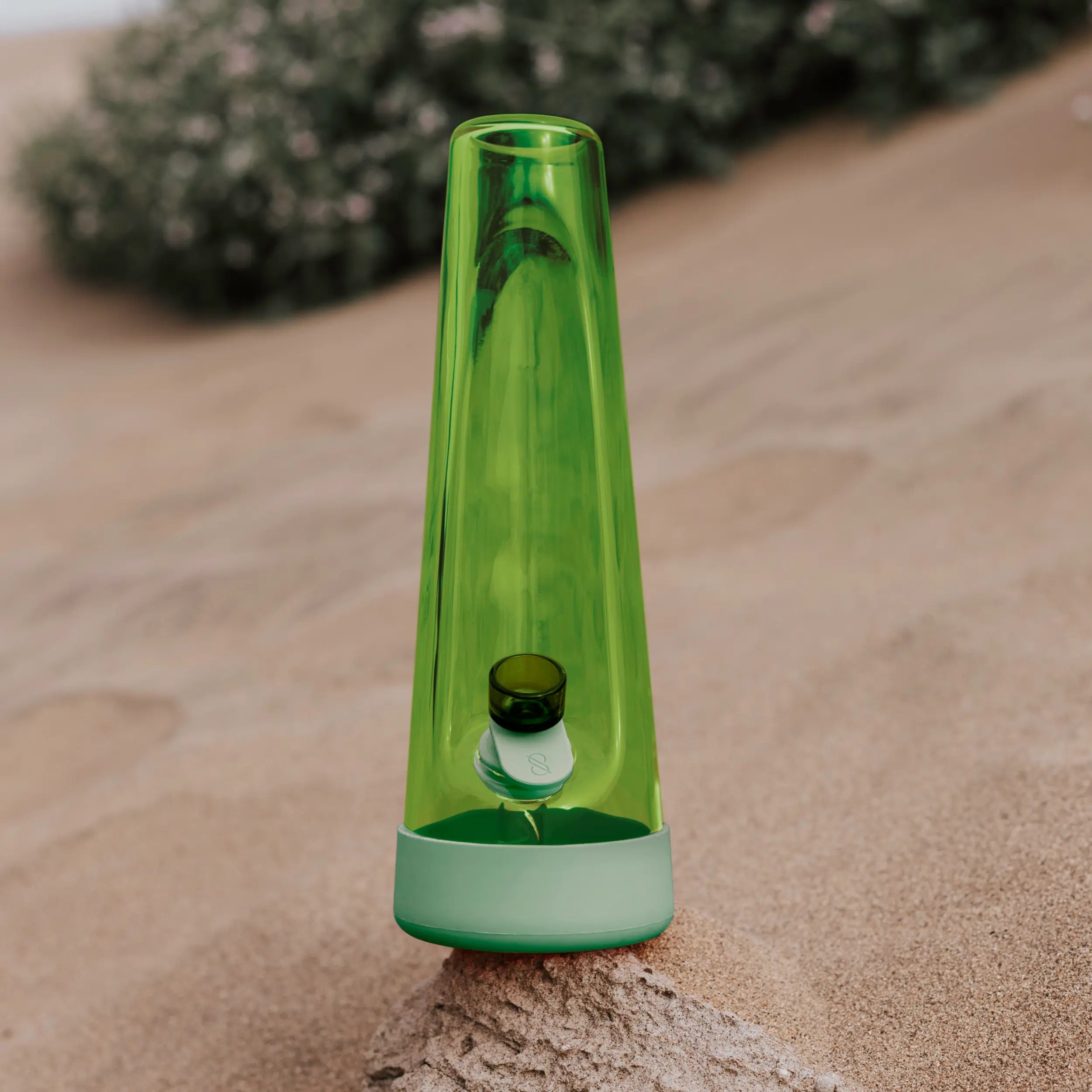 Session Goods Designer Series Grass Bong standing on sand with a blurred background, featuring sleek green glass and soft green silicone