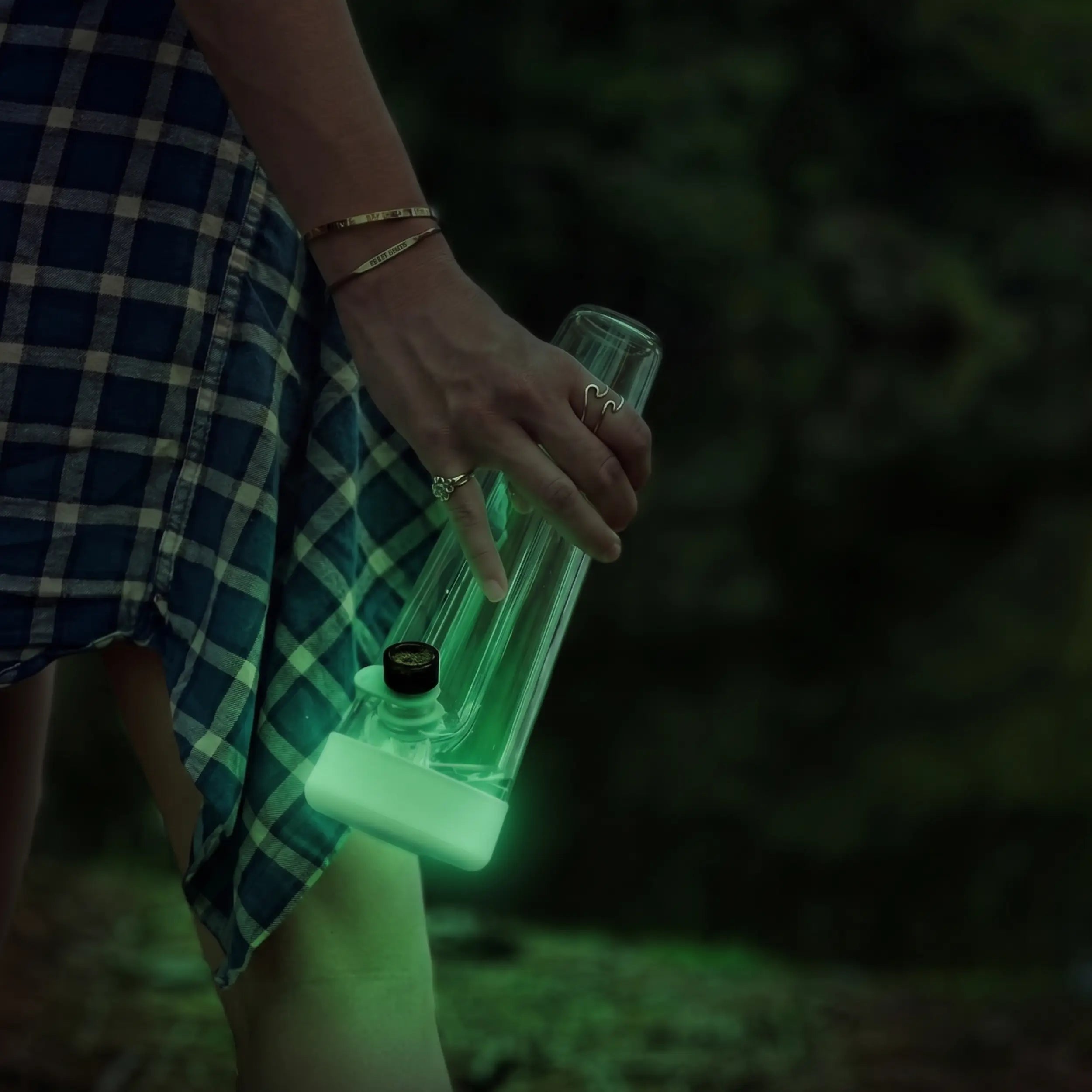 Person walking on an outdoor night trail holding the Session Goods glow-in-the-dark glass bong, featuring a luminescent lime green silicone base illuminating the dark.