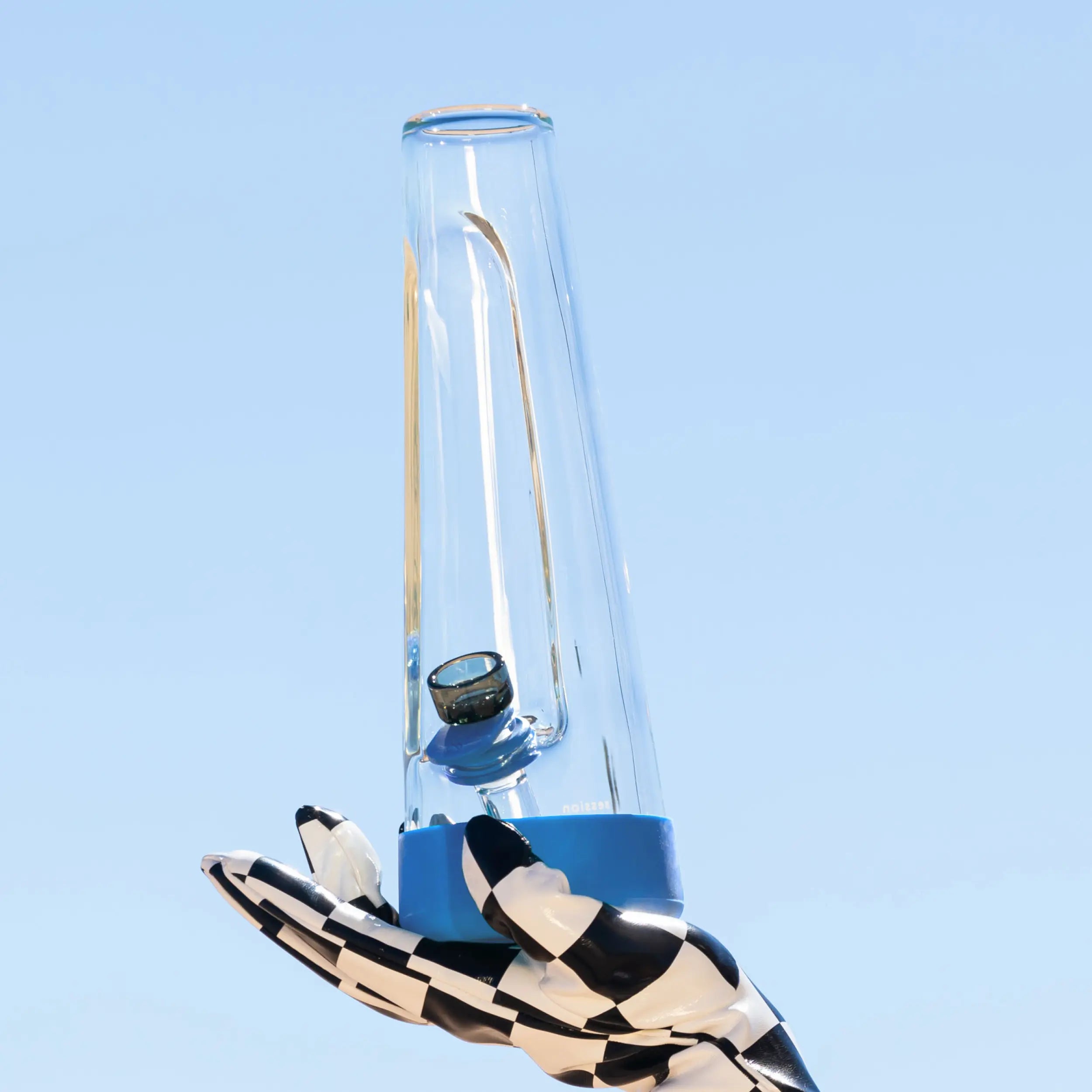 A striking, artistic image of a hand in a checkered glove holding the Cyan Blue Session Bong up against a clear, bright blue sky, highlighting the lucid colorway.