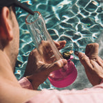 A sun-drenched lifestyle shot of someone using the Magenta Pink Session Bong by a sparkling blue pool, emphasizing the heat-resistant silicone and summer vibe.