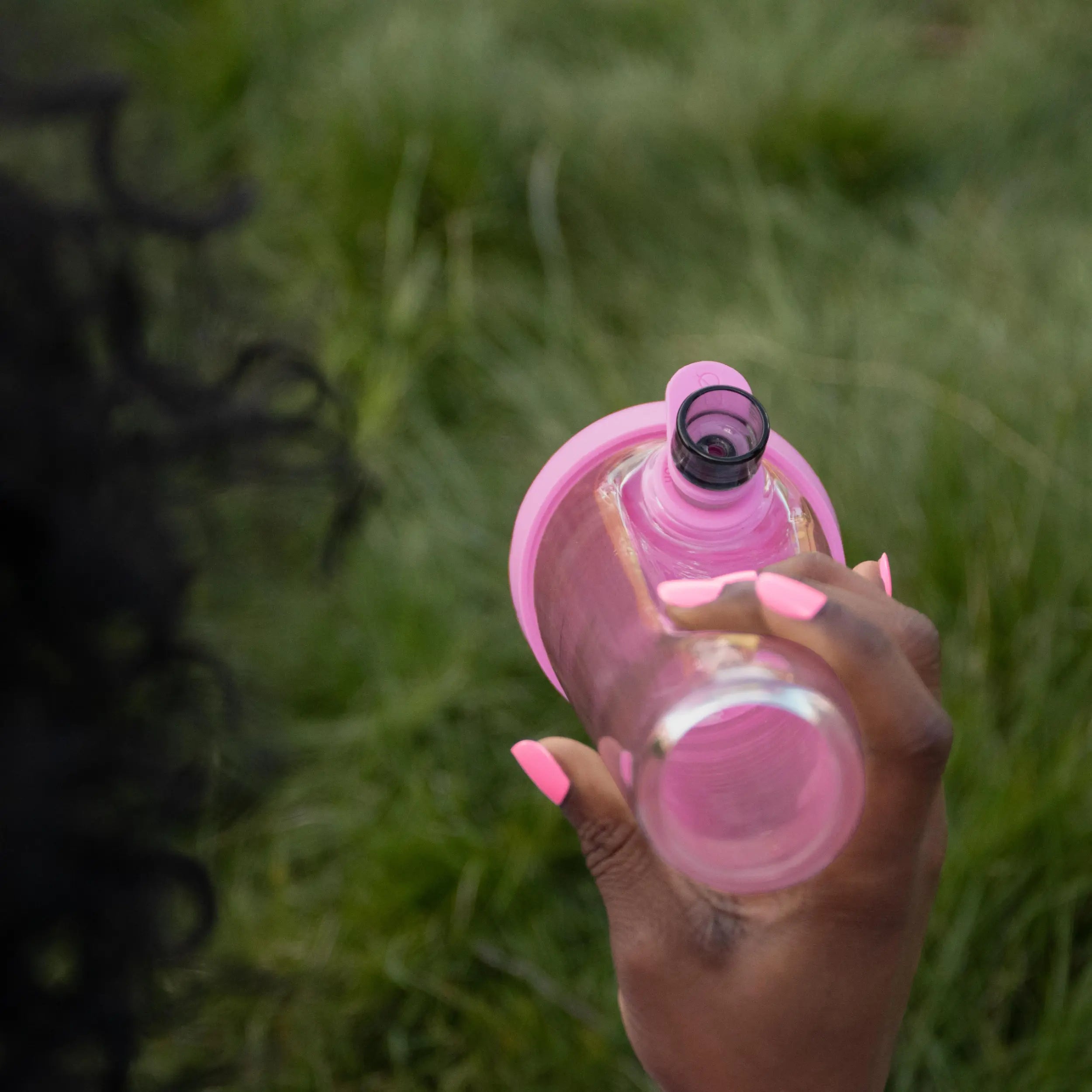 An overhead lifestyle view of a person with pink manicured nails holding the Magenta Pink Session Bong against a soft green grass background.
