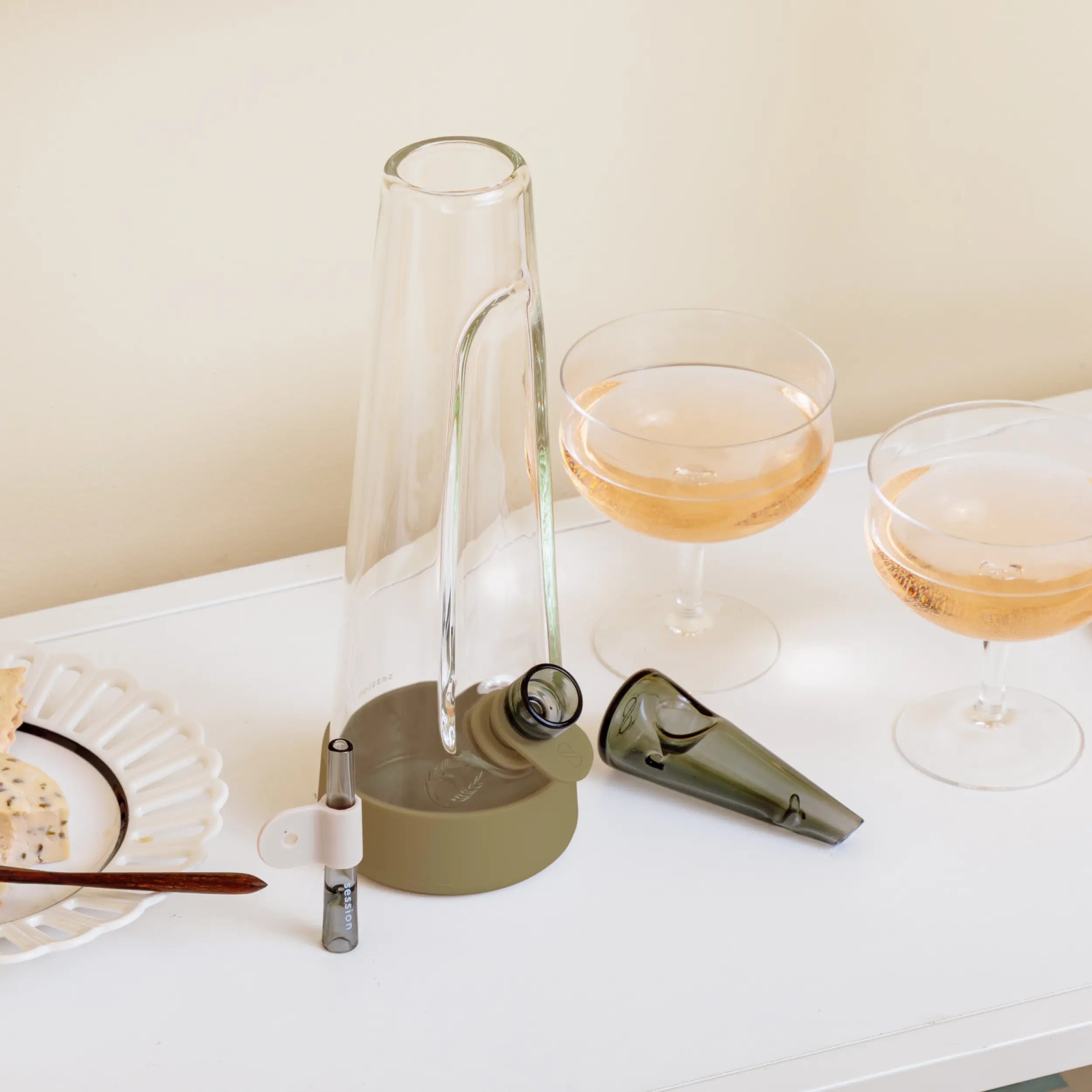 A lifestyle arrangement on a white table featuring a 10 inch, olive green Session bong, two glasses of rosé wine, and a small plate of snacks. The bong's bowl and downstem accessories are shown detached next to the main glass piece.