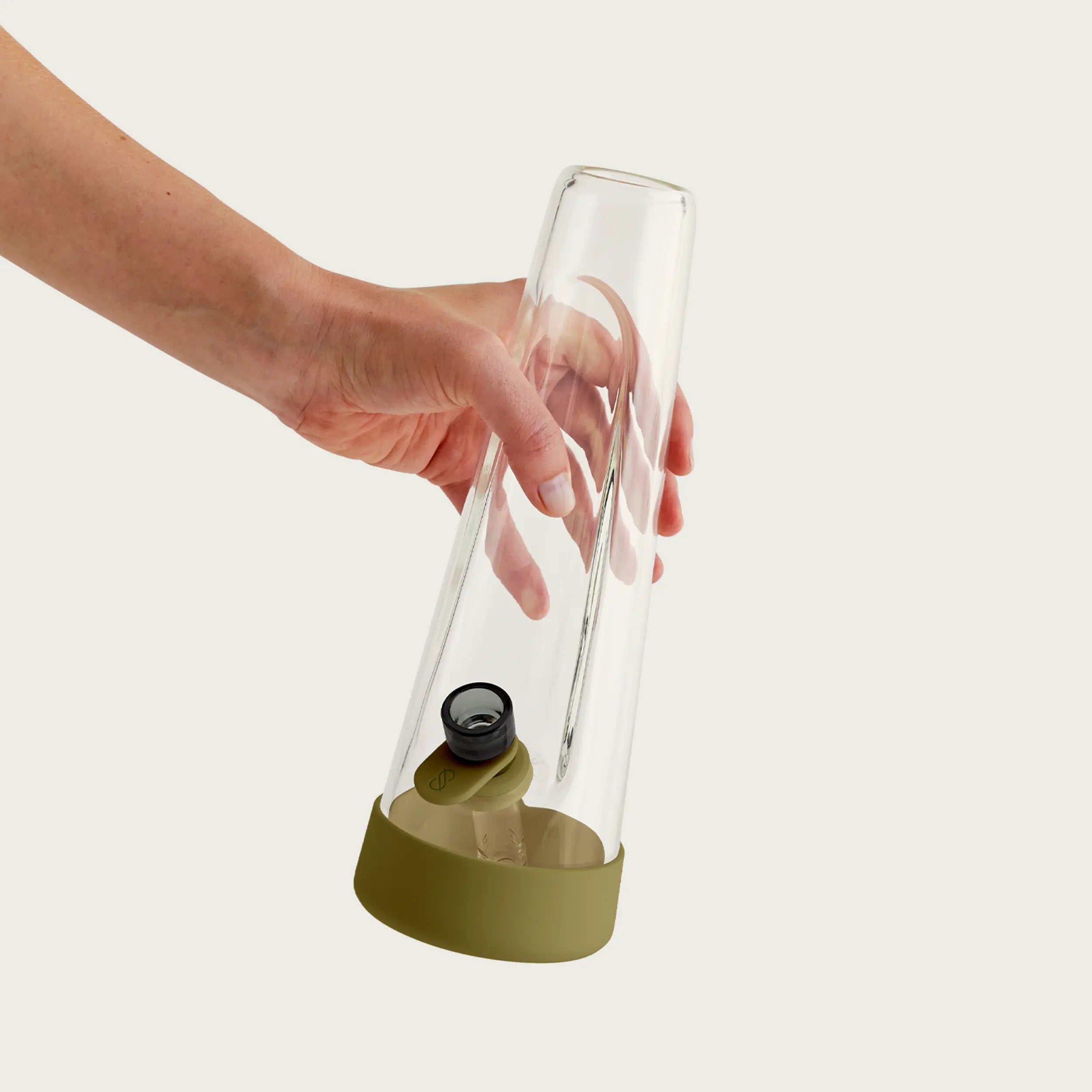 A person’s hand holds a 10 inch Session Goods glass bong with olive green silicone accents. The tapered, modern glass design is shown against a plain light gray background, highlighting its portable size and ergonomic shape.
