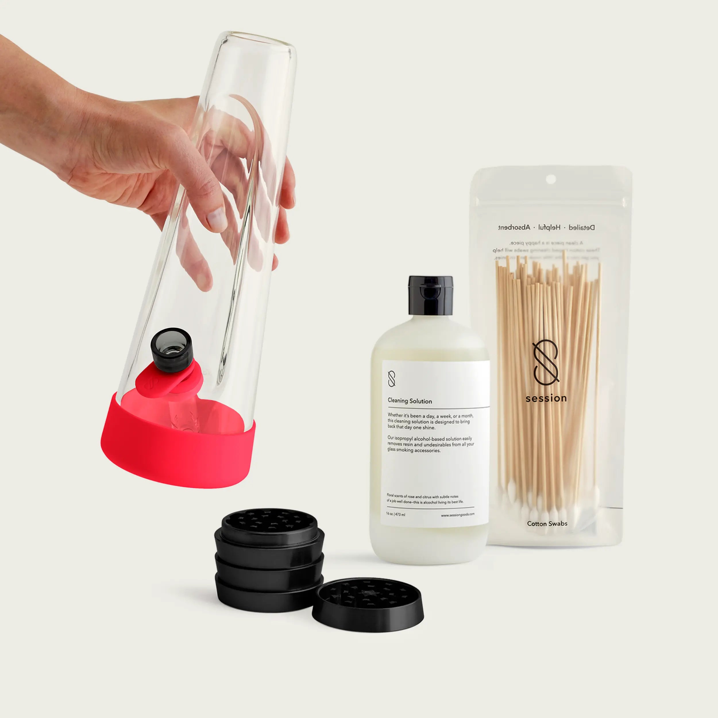 A hand holding a clear glass Session Goods bong with a cherry red silicone base and tab, displayed alongside a black multi-tier grinder, a bottle of Session cleaning solution, and a pouch of wooden cotton swabs.