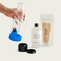 A hand holds a clear glass Session Bong with Cyan Blue silicone accents alongside the essential starter components: a matte black precision grinder, a bottle of residue-free cleaning solution, and a pack of detailed cotton swabs.