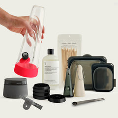A curated collection of modern, minimalist smoking accessories arranged on a clean, off-white background. A hand is lifting a clear, conical glass bong featuring a bright Cherry Red silicone base and bowl attachment. Scattered around the bong are a bottle of Session cleaning solution, a bag of wooden cleaning swabs, multiple black and charcoal silicone cleaning and storage pouches, a disassembled matte black multi-piece grinder, a dark grey stash jar, two cone-shaped portable glass pipes, a small one-hitter