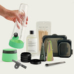 The Session Goods Complete Bundle arranged on a light background. A hand is lifting a clear glass bong featuring a glow lime green silicone base. Arranged below are a matching lime green stash jar with a dark lid, a disassembled black metal grinder, a small grey one-hitter, a bottle of cleaning solution, wooden detailing sticks, a glass pipe with a swirled silicone sleeve, three dark translucent silicone cleaning pouches in varying sizes, and a dark metal measuring spoon.