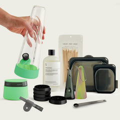 The Session Goods Complete Bundle arranged on a light background. A hand is lifting a clear glass bong featuring a glow lime green silicone base. Arranged below are a matching lime green stash jar with a dark lid, a disassembled black metal grinder, a small grey one-hitter, a bottle of cleaning solution, wooden detailing sticks, a glass pipe with a swirled silicone sleeve, three dark translucent silicone cleaning pouches in varying sizes, and a dark metal measuring spoon.