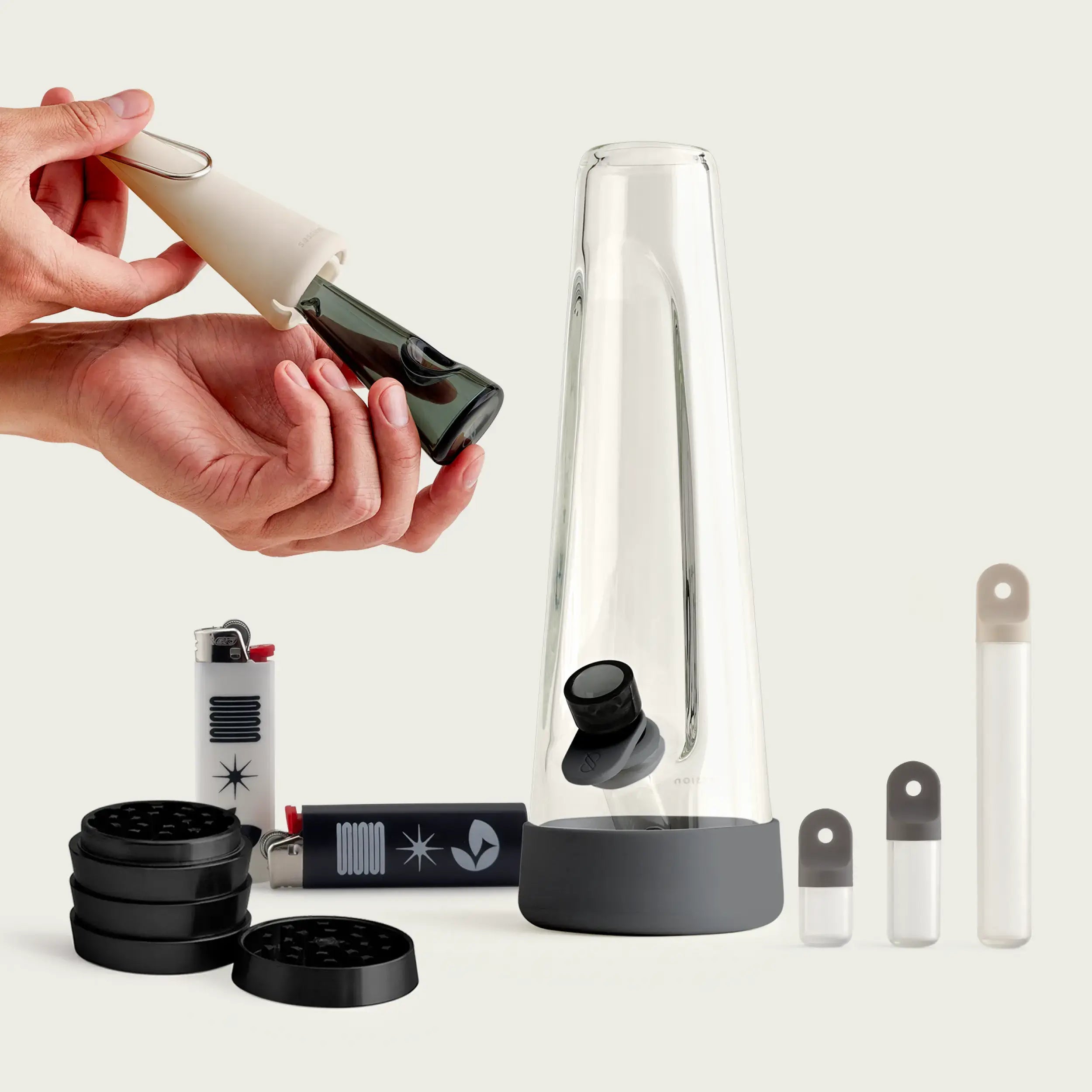 A full Black Friday bundle from Session Goods, centered around the Session Bong (a sleek, clear glass water pipe with a grey base). The bundle also includes the Session Pipe being held by a pair of hands (a small glass pipe with a cream-colored silicone sleeve/cover), a stack of three black Session Grinders, and two branded lighters (one white, one black). To the right are three small, clear containers with white silicone caps, which appear to be storage tubes.