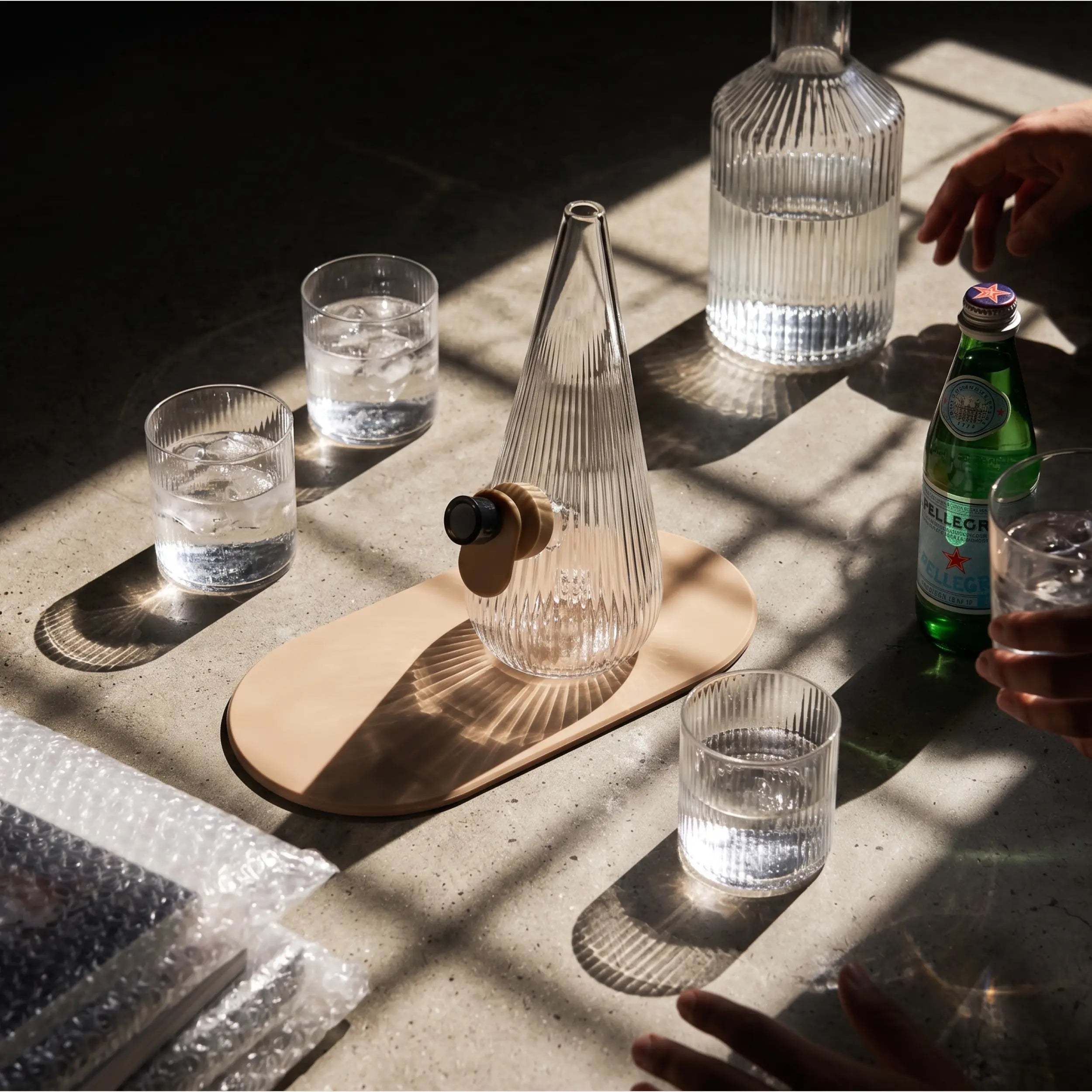Lifestyle shot of the Session Goods ribbed glass Drop Bubbler in sand on a sunlit table surrounded by drinking glasses, a carafe, and San Pellegrino bottle.