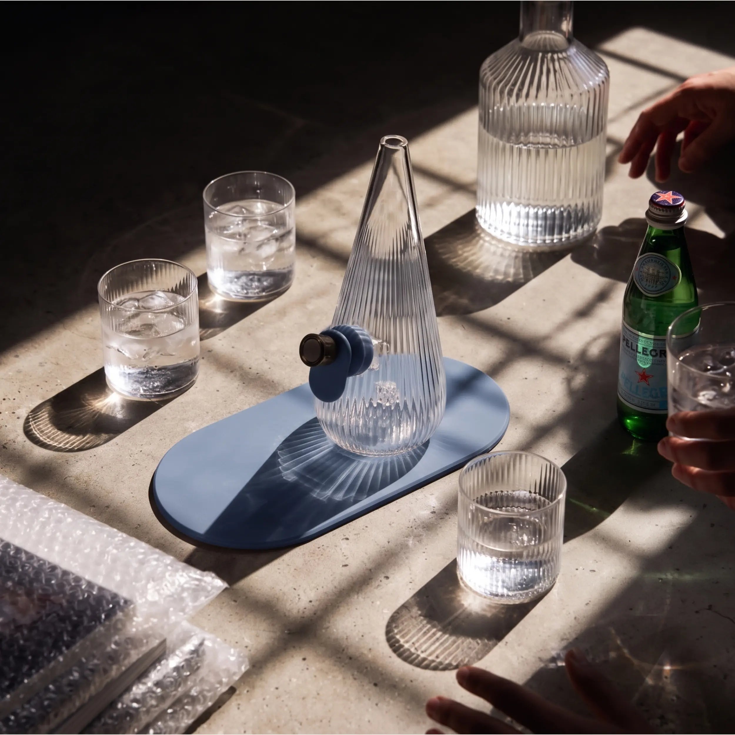 Lifestyle shot of the Session Goods ribbed glass Drop Bubbler in slate blue on a sunlit table surrounded by drinking glasses, a carafe, and San Pellegrino bottle.