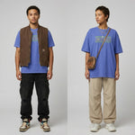 A studio shot of two models showcasing a blue graphic t-shirt against a plain white background. On the left, a young man wears the blue tee under a brown zip-up work vest, paired with distressed black cargo pants and multi-colored high-top sneakers. On the right, a young woman wears the same blue t-shirt featuring the neon green and yellow text "CAMP DO NOTHING", styled with loose beige pants, chunky white sneakers, layered silver necklaces, and a small brown leather crossbody bag.