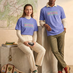 A lifestyle photo of a man and woman in an indoor setting with a rustic, exploration theme. Both wear the periwinkle 'CAMP DO NOTHING' t-shirt. The woman, sitting with a camera, layers hers over a cream turtleneck. The man, standing next to her and leaning, wears his over a cream knit polo collar. A large map of 'Pinewood Trails' and stacked books are in the background