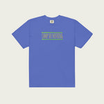 A flat-lay view of a short-sleeved, garment-dyed periwinkle blue t-shirt on a pale tan background. The front of the shirt features the central orange-pink 'CAMP DO NOTHING' screen-print graphic with its green zigzag borders.