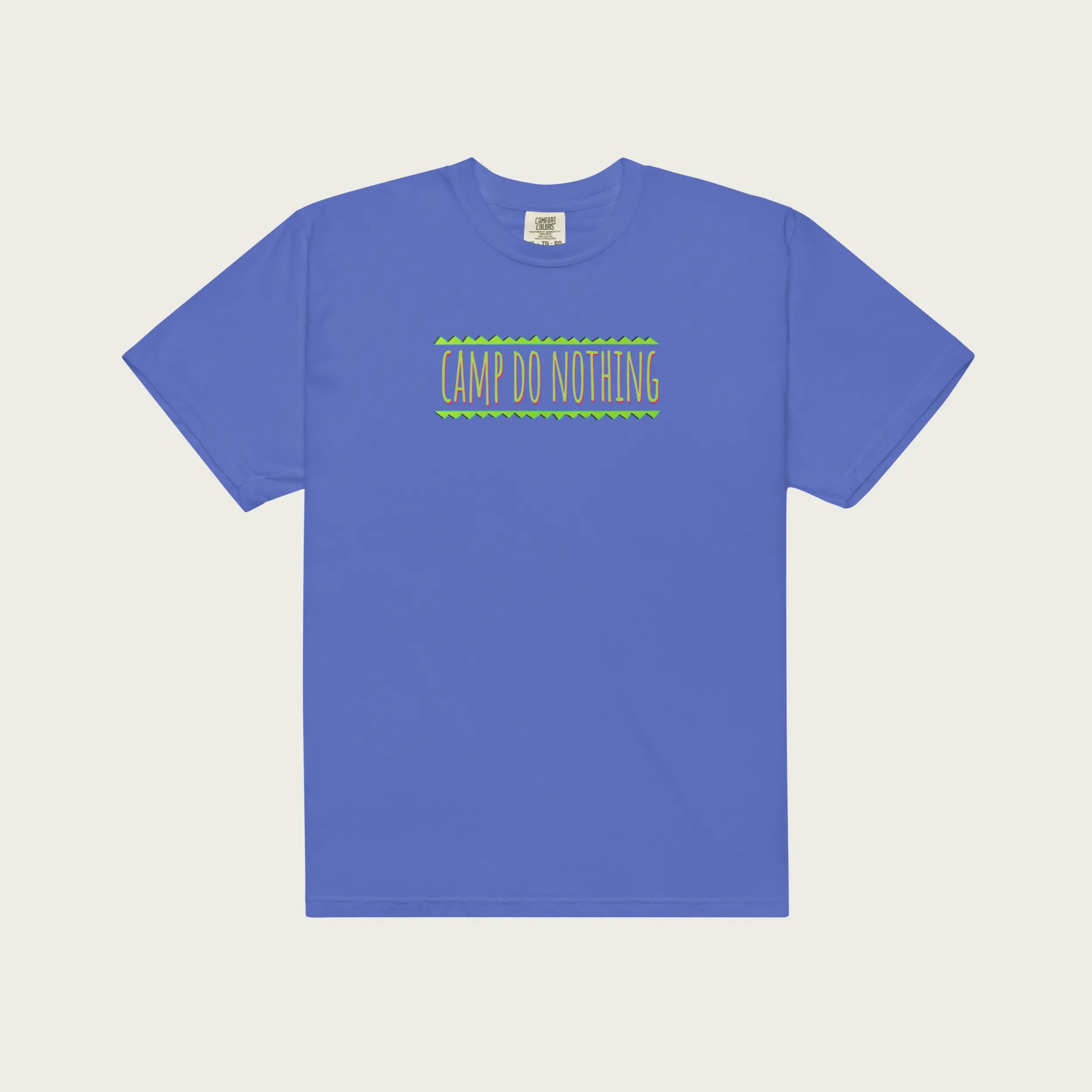 A flat-lay view of a short-sleeved, garment-dyed periwinkle blue t-shirt on a pale tan background. The front of the shirt features the central orange-pink 'CAMP DO NOTHING' screen-print graphic with its green zigzag borders.