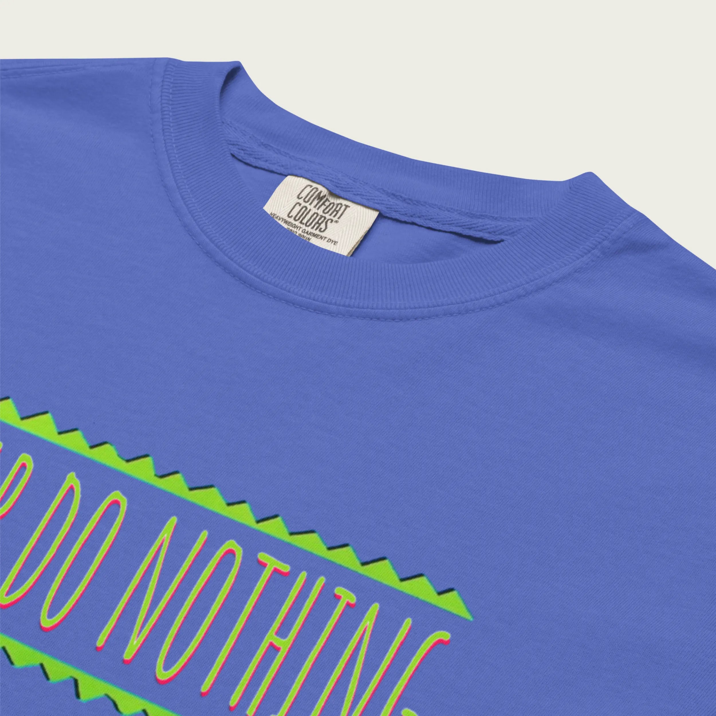 A close-up photograph of the ribbed knit neckline of a periwinkle blue t-shirt. A woven 'Comfort Colors' tag is visible inside the collar, with fine text. A corner of the green zigzag graphic is visible below the neckline