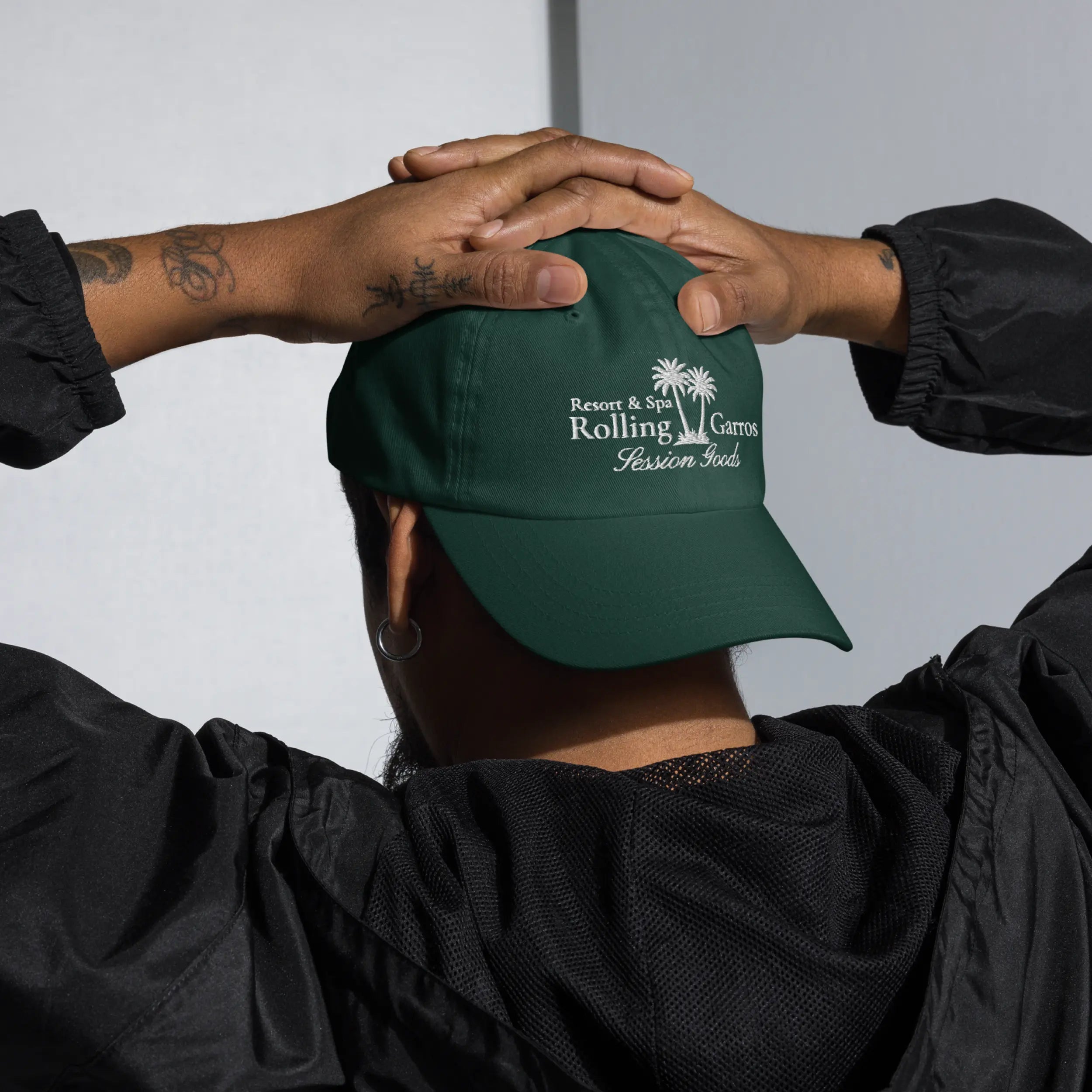 Person wearing green Session Goods Rolling Garros dad hat with embroidered palm tree logo, hands resting on head.