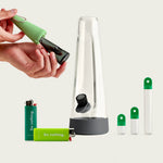 A lifestyle product shot of the Session Goods Shamrock St. Paddy's Day Bundle featuring a clear glass bong with a charcoal gray silicone base, a smoke-tinted glass pipe being inserted into a celery green silicone sleeve, three glass stash tubes with vibrant green caps, and two "Do Nothing" branded BIC lighters in lime and forest green against a clean white background.
