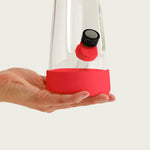A hand holding the bottom of a clear glass Session Goods bong, highlighting the installed cherry red silicone protective base and matching silicone bowl tab.