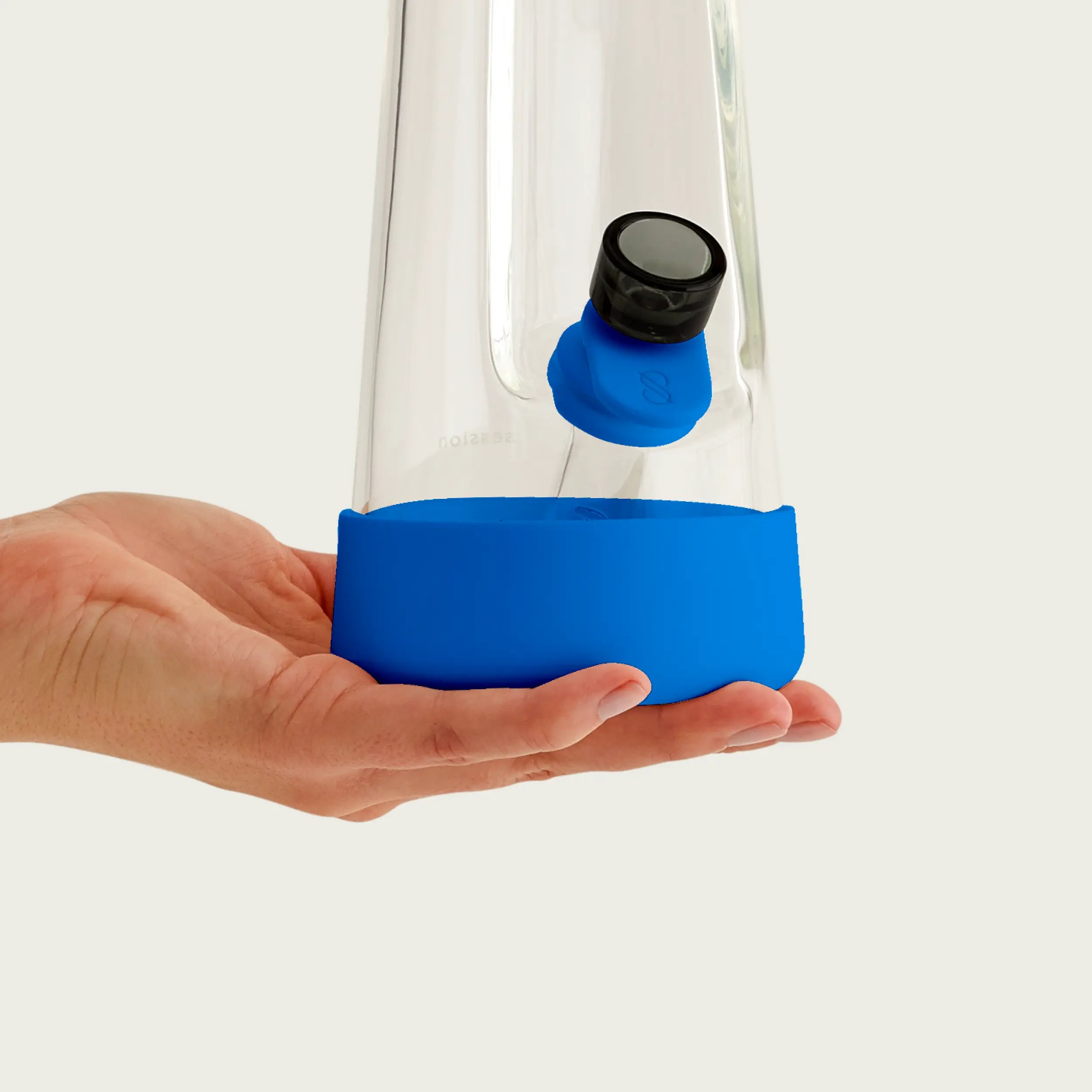 A hand holding the base of a clear glass Session Bong fitted with the Cyan Blue silicone accessory set. The image highlights the crisp blue protective footer and the coordinating pull tab for a clean, modern look.